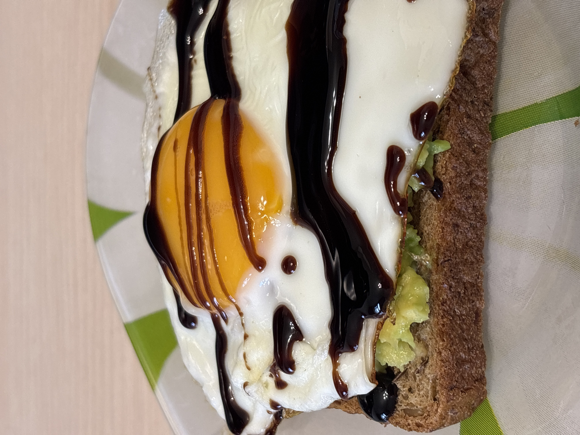 The image shows a close-up of a partially eaten open-faced sandwich on a plate with green and white stripes. The sandwich consists of a slice of dark bread topped with mashed avocado, a fried egg, and dark drizzled sauce. The plate sits on a surface with a light-colored, possibly wooden, pattern. There is no evidence of a location in Rusanovo, Russia.