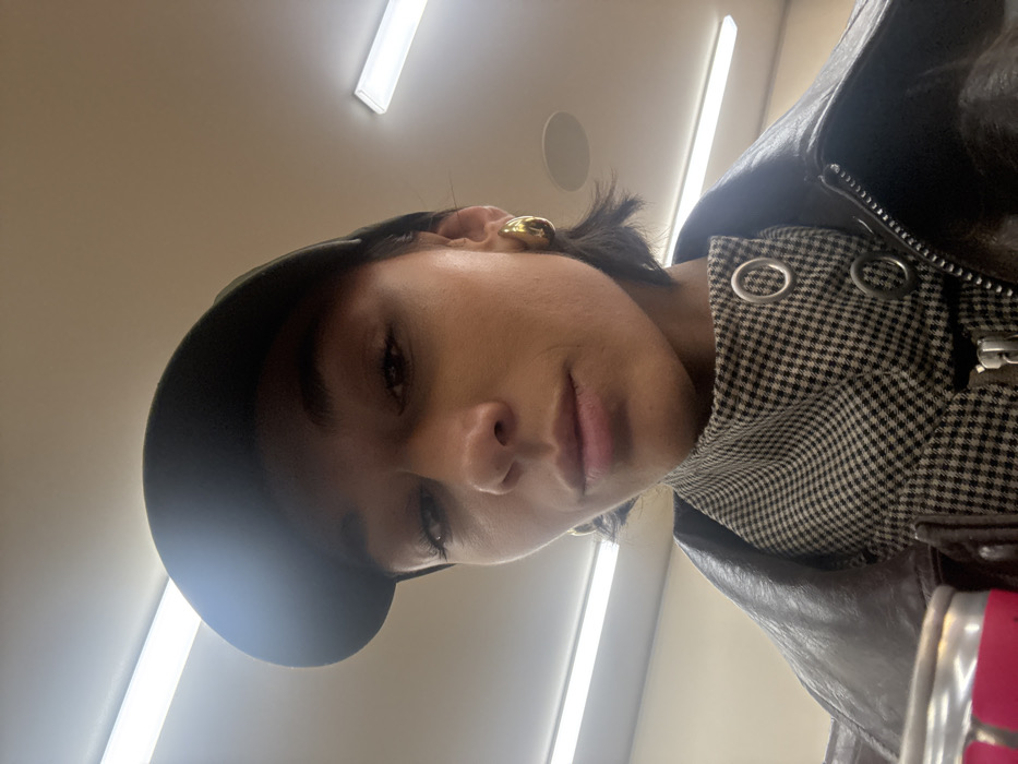 A person wearing a dark cap, leather jacket, and a patterned shirt or jacket underneath. The person has dark hair, and a gold hoop earring is visible. The picture is taken from a low angle, looking up at the person, with fluorescent lights in the background. The lighting is bright, and the image is slightly rotated.