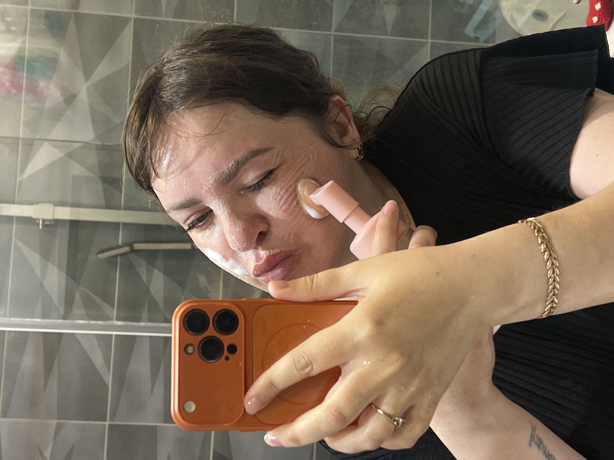 The image captures a close-up, mirrored selfie of an adult individual performing a skincare routine in a bathroom. The person has dark brown hair, which appears slightly damp, and a white or cream-colored product is actively applied to their face, visible on the forehead, cheeks, and chin in distinct streaks. The individual's lips are pursed in a pouting expression, and their eyes are partially closed.
In the left foreground, the individual's left hand holds an orange-cased smartphone, equipped with a triple-lens camera system, indicating it is being used to capture the reflected image. A gold ring with a small, clear stone is present on the ring finger of the left hand. The right hand, in the center-right of the frame, holds a light pink skincare tool with a rounded, applicator tip that is in direct contact with the right cheek, applying the facial product. A gold-colored chain bracelet is visible on the individual's right wrist. A faint, illegible tattoo is partially visible on the inner left forearm. The individual is wearing a black, ribbed, short-sleeved top.
The background, reflected in the mirror, consists of dark grey square wall tiles featuring a subtle, lighter grey geometric pattern. A vertical metallic frame, characteristic of a shower enclosure, is prominent on the left side of the reflection, alongside a white wall-mounted fixture. The upper right background shows blurred reflections of various objects, and a red and white polka-dotted item is faintly visible in the lower right. The specific location in the city of Noviy, Russia, cannot be determined from the visual information in the image.
