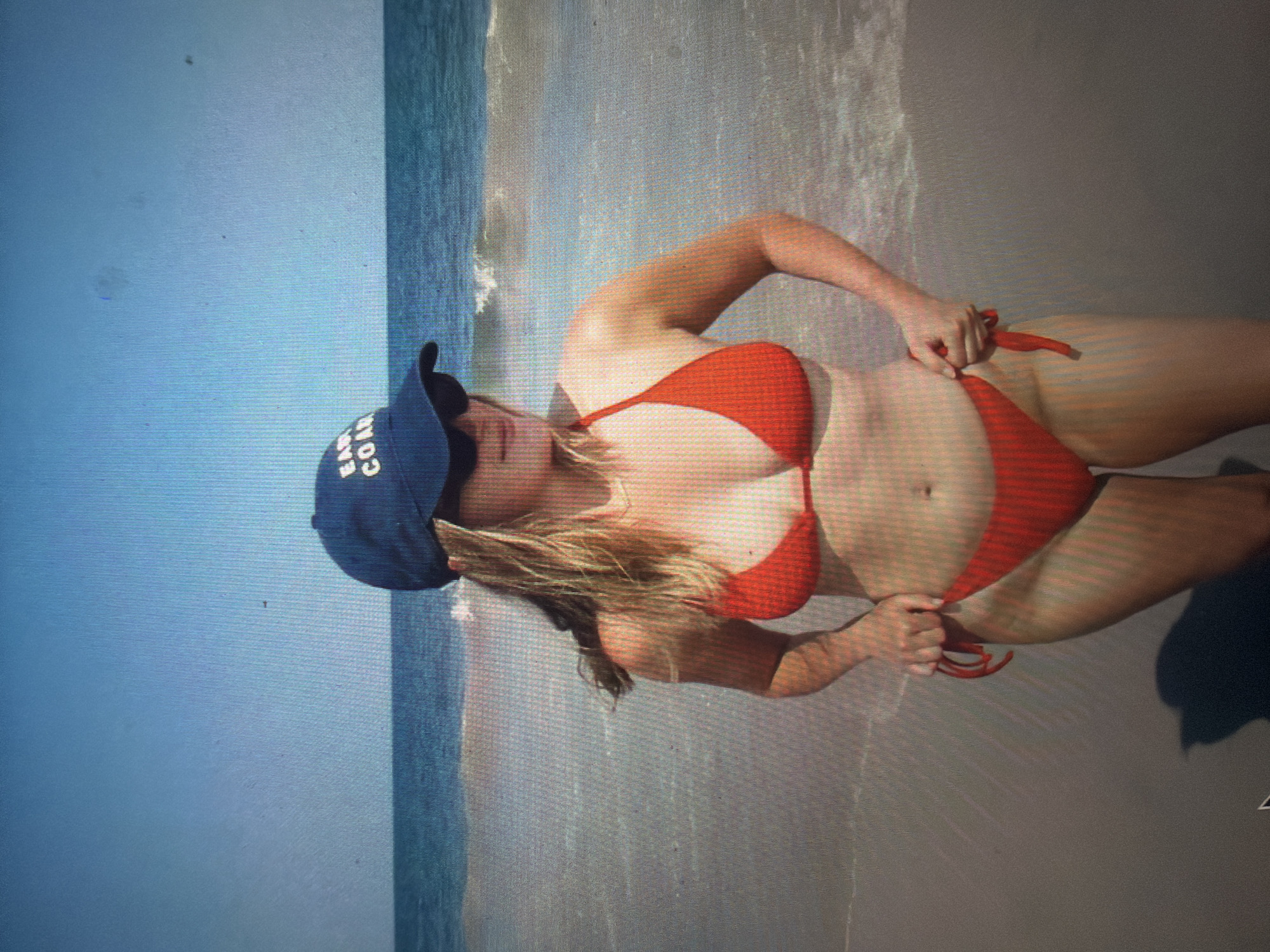 A young adult female stands on a sandy beach at the ocean's edge under a clear blue sky. She wears a red string bikini, dark sunglasses, and a dark blue baseball cap with white lettering that reads "EAST COAS" (likely "EAST COAST"). Her long, light-colored hair extends over her shoulders. She is positioned slightly angled to the viewer's right, looking downwards, with both hands holding or adjusting the sides of her bikini bottom. Gentle waves break on the wet sand behind her. The environment is bright and sunny. There are no discernible landmarks, architectural features, or specific environmental cues within the image that indicate the location is in the city of Shumen, Bulgaria; the scene depicts a generic coastal setting.