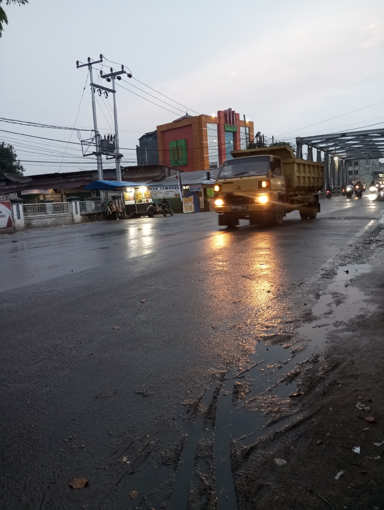 The image captures a street scene in Air Molek, Indonesia, likely during twilight or overcast conditions given the low light and artificial lighting illumination. A yellow dump truck is the prominent subject, traveling on a wet asphalt road. The road surface displays reflections from the truck's headlights and other light sources, indicating recent rain or ongoing moisture. Tire tracks and puddles of water are visible on the road, suggesting poor drainage or the aftermath of rainfall.
On the left side of the road, a low-rise building with a white fence in front of it is partially visible. There is a small food stall with a blue awning and lights illuminating its wares. Two individuals are seated on a bench in front of the stall. Several power poles with electrical transformers and wires are prominent in the background, typical of urban infrastructure.
Further down the road, a bridge structure is visible, with more vehicles, including motorcycles, appearing to cross it. The architecture of a building in the distance, with orange and green sections, suggests a commercial or institutional establishment. The overall atmosphere is that of a moderately busy street with ongoing traffic and pedestrian activity.
