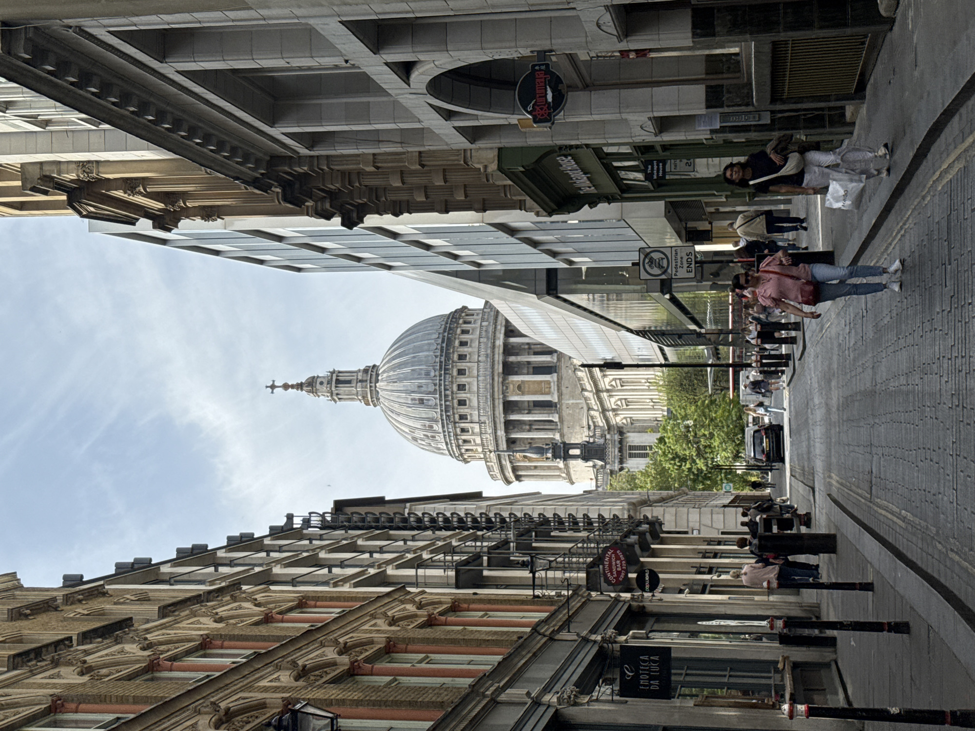 The image shows a view down a narrow street in London, United Kingdom, with St. Paul's Cathedral in the background. The street is lined with buildings of varying architectural styles, from older, ornate facades to modern glass-fronted structures. Pedestrians are walking along the street, and street signs and business signs are visible. The street is paved with cobblestones.