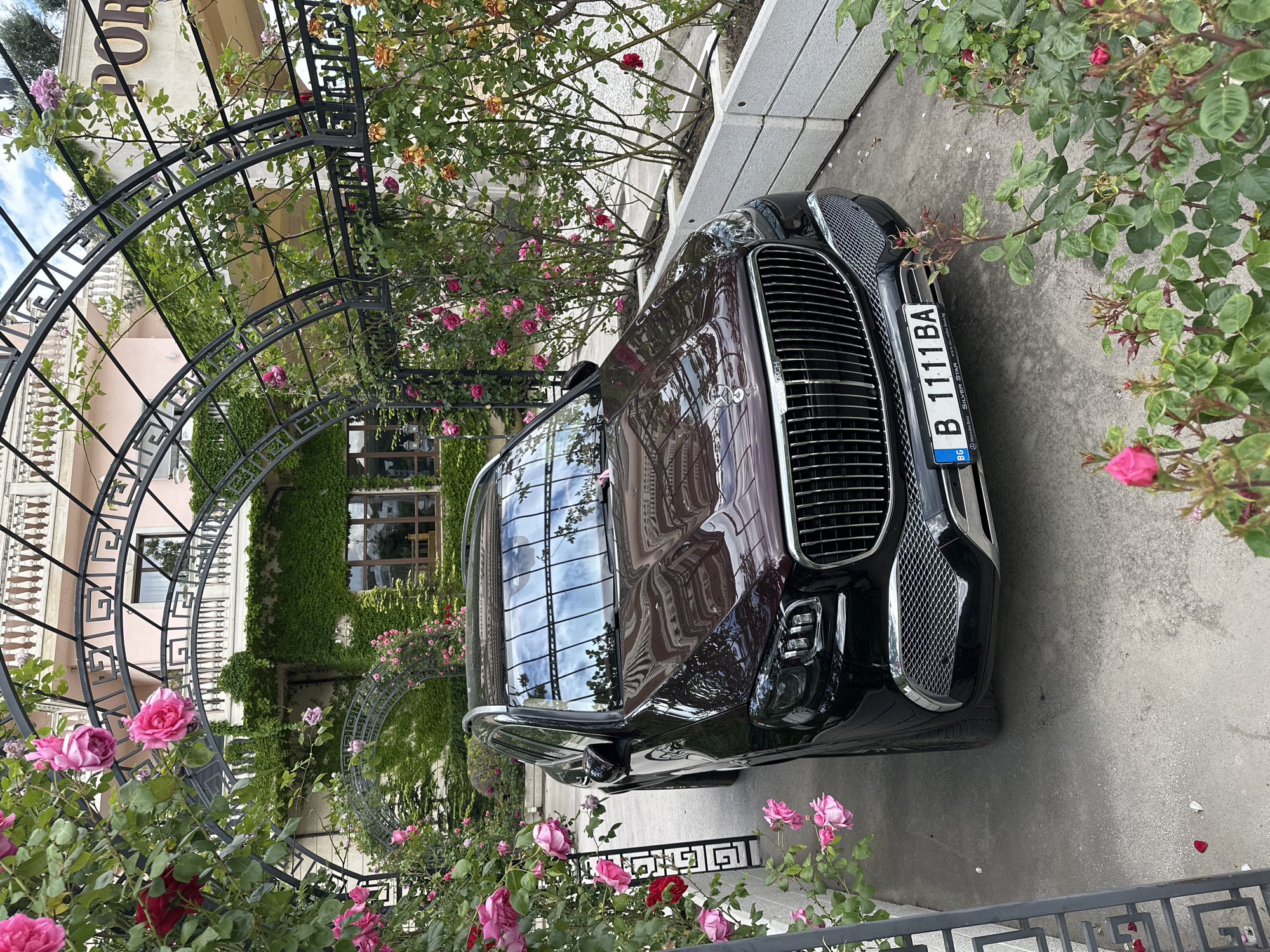 The image captures a dark burgundy Mercedes-Benz SUV parked on a gray concrete surface. The car has a prominent chrome grill and a Bulgarian license plate reading "B 1111 BA". The surrounding environment includes an elaborate metal pergola covered with climbing roses in shades of pink and red. The architectural style of the building in the background is light-colored with ornate detailing. The scene is located in Varna, Bulgaria.