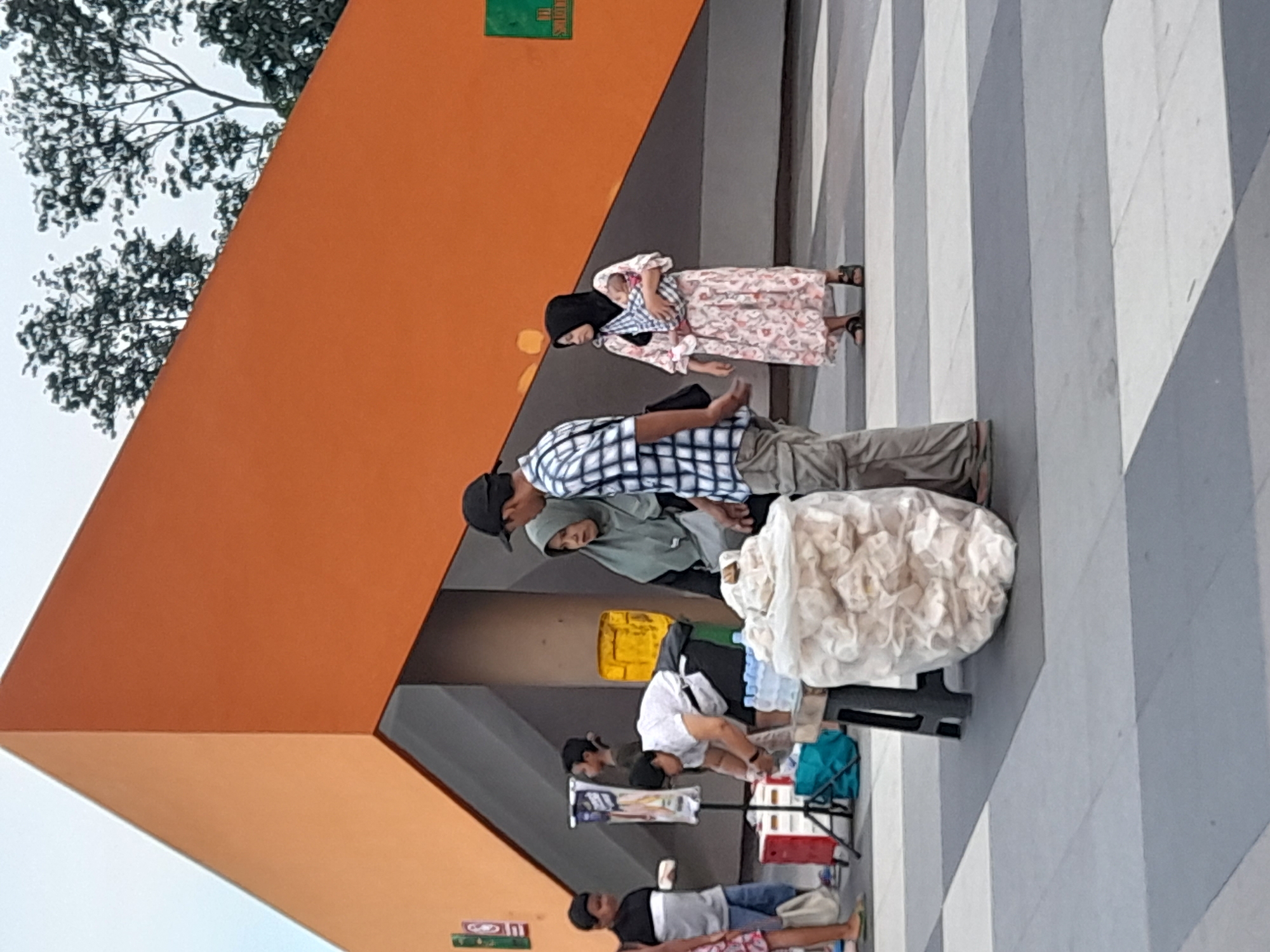 A rotated eye-level shot captures an outdoor scene, potentially in a public area. Several individuals are present, with a woman wearing a floral dress and hijab visible near an unidentified man.
Further down, a group of people appears to be involved in some kind of activity or transaction. A large, rounded, white object sits on a black stand or frame. Various objects are arranged around this area, including what appears to be a yellow container, colorful crates, and assorted goods or merchandise.
The setting includes a distinctive orange structure with a grey awning or overhang. The ground features a pattern of alternating light and dark grey stripes or tiles. A tree with dense foliage is visible in the background.
The location is identified as the city of Sidoarjo, Indonesia.