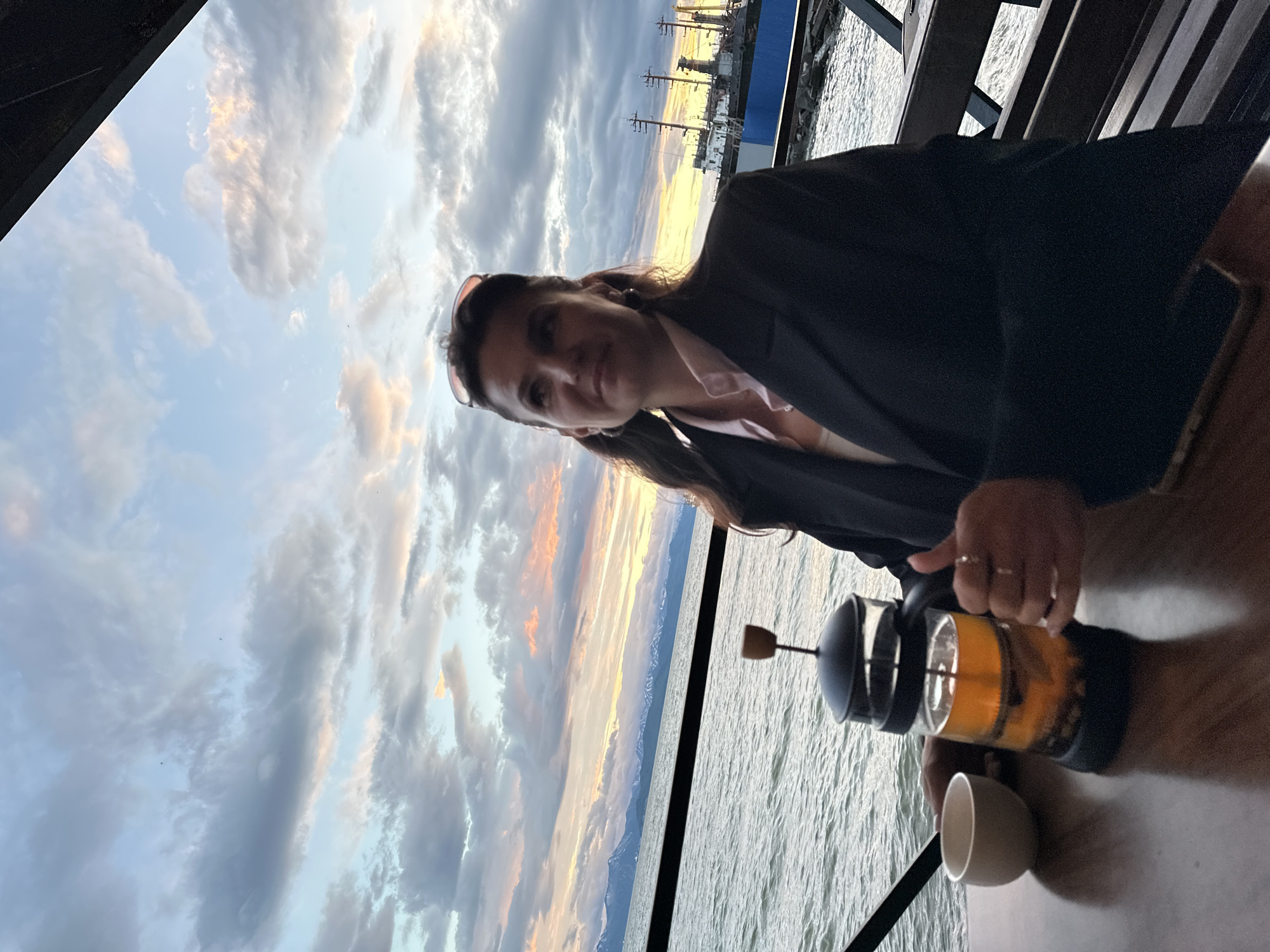 A woman with long, dark hair is seated at a table by a body of water, likely during sunset or sunrise. She is wearing a black blazer over a pink collared shirt and a light-colored undershirt. Sunglasses are perched on her head. In front of her is a glass French press filled with amber liquid, presumably tea, and a small white teacup. Her hands are on the table, one near the French press and the other resting on her lap. In the background, the water stretches towards a distant coastline with snow-capped mountains. Several ships are docked in the harbor, and the sky is filled with dramatic clouds tinged with orange and pink hues. The scene suggests a tranquil moment by the water, possibly in Petropavlovsk-Kamchatskiy, Russia, given the maritime setting and mountainous backdrop typical of the region.