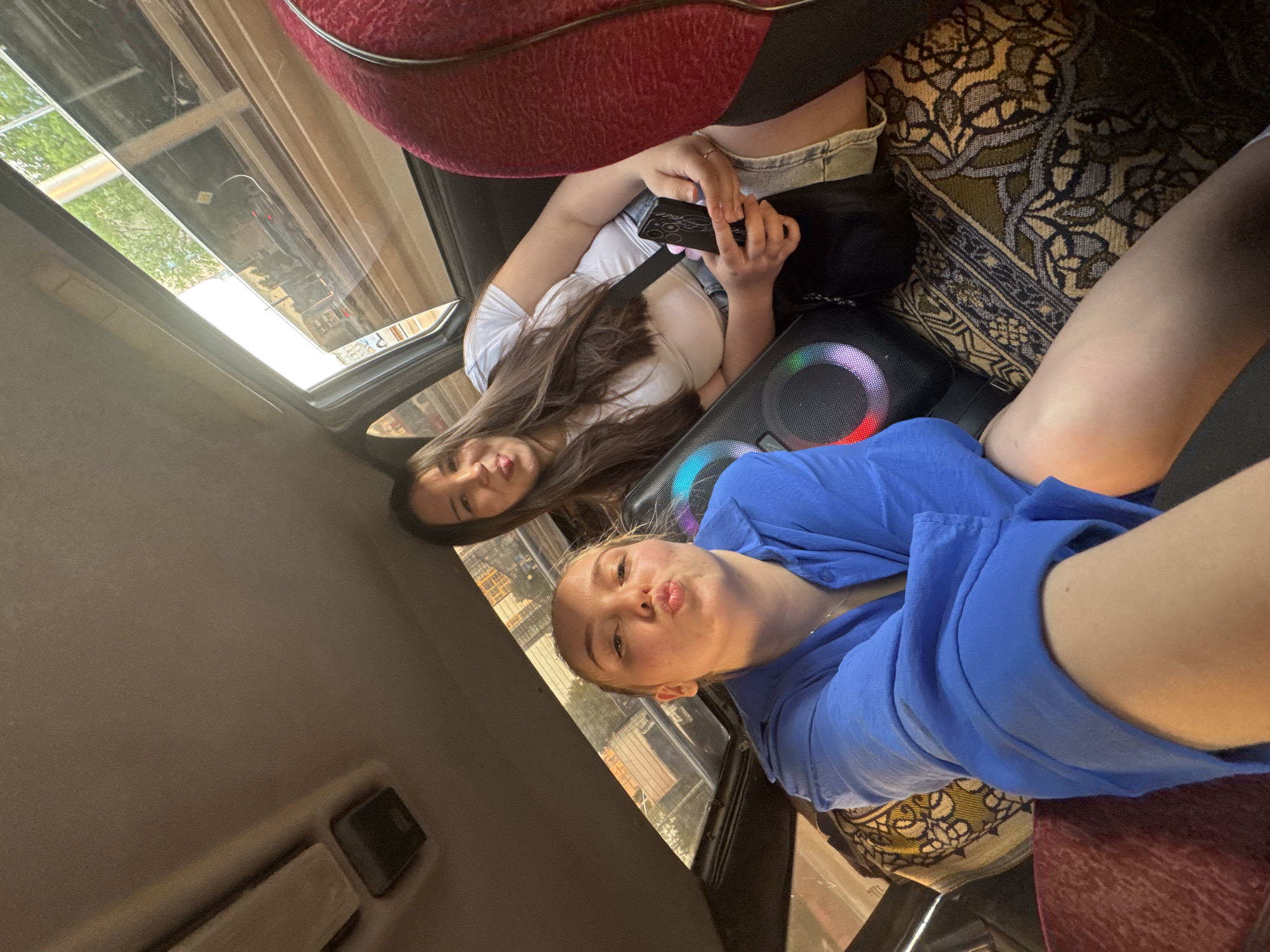 Two women are inside what appears to be a vehicle, possibly a bus or a specialized transportation service, with a cityscape visible through the window. One woman is wearing a blue dress and is closer to the camera, making a kissing face. The other woman is seated in the back, wearing a white shirt and denim shorts, holding a smartphone. There is a black speaker with colored lights between them. The seat upholstery is patterned. The photo's perspective is angled upwards, capturing the open sunroof and a glimpse of the city outside. The location is likely Perm, Russia, based on the visible urban architecture.