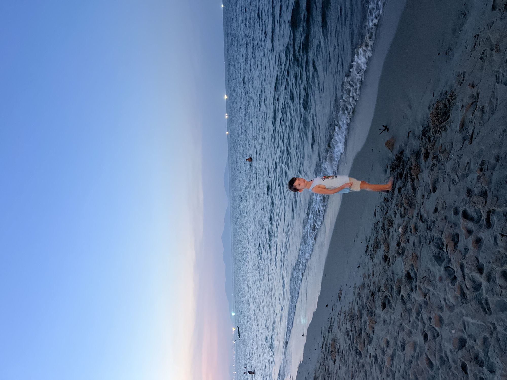 A young boy stands on a sandy beach in Hoi An, Vietnam, facing the ocean during twilight. The sky displays a gradient of soft blues and pinks, transitioning to a deeper blue as it reaches higher. The sea is calm with gentle waves lapping the shore. Distant lights dot the horizon, suggesting a coastal city or boats at sea. In the background, mountains are faintly visible, silhouetted against the dusk sky. Other beachgoers and a small boat are present in the water. The sand is textured with footprints and small mounds, indicative of a well-used beach. The boy is wearing a white tank top and tan shorts, and appears to be looking towards the camera.