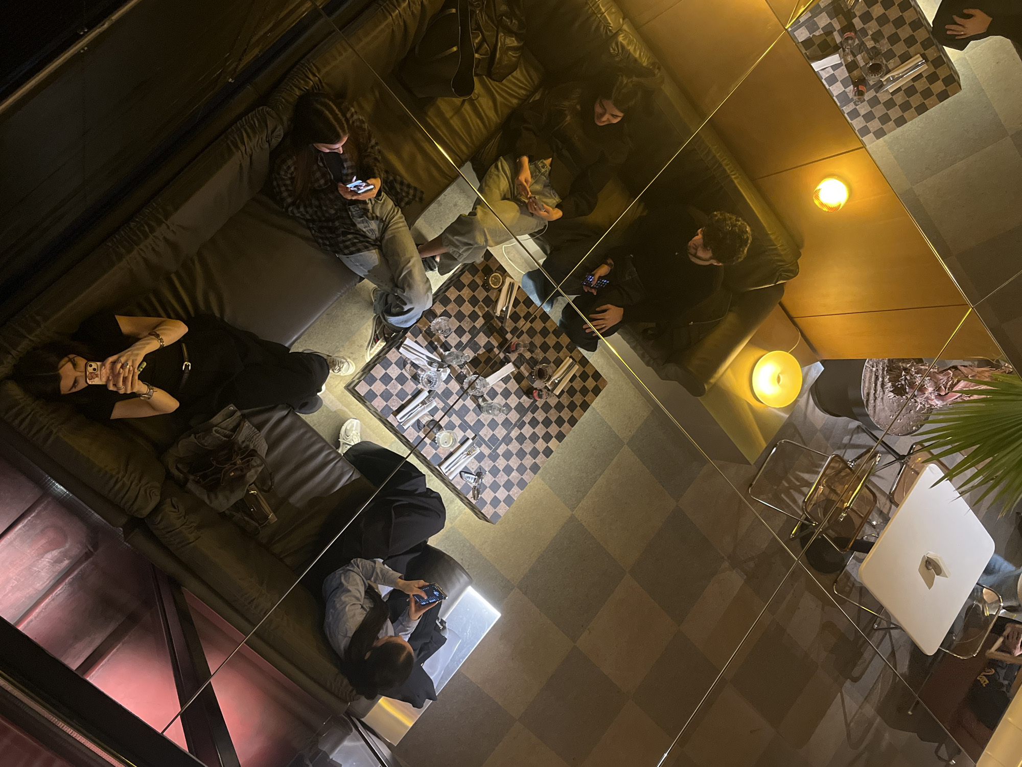 A high-angle, medium shot depicts a group of people in an indoor space, possibly Yerevan, Armenia, given the style. Six individuals are visible, seated on plush, dark couches arranged to create a lounge-like atmosphere. Each person appears engaged with their mobile phones. A table with a checkered top is set with glassware and cutlery, suggesting a meal setting. The floor is also tiled in a checkered pattern. The walls are lined with reflective surfaces, creating a mirrored effect that complicates the perception of the space's size and layout. Lighting is provided by ceiling fixtures, as well as lamps. The overall ambiance is dim and intimate.