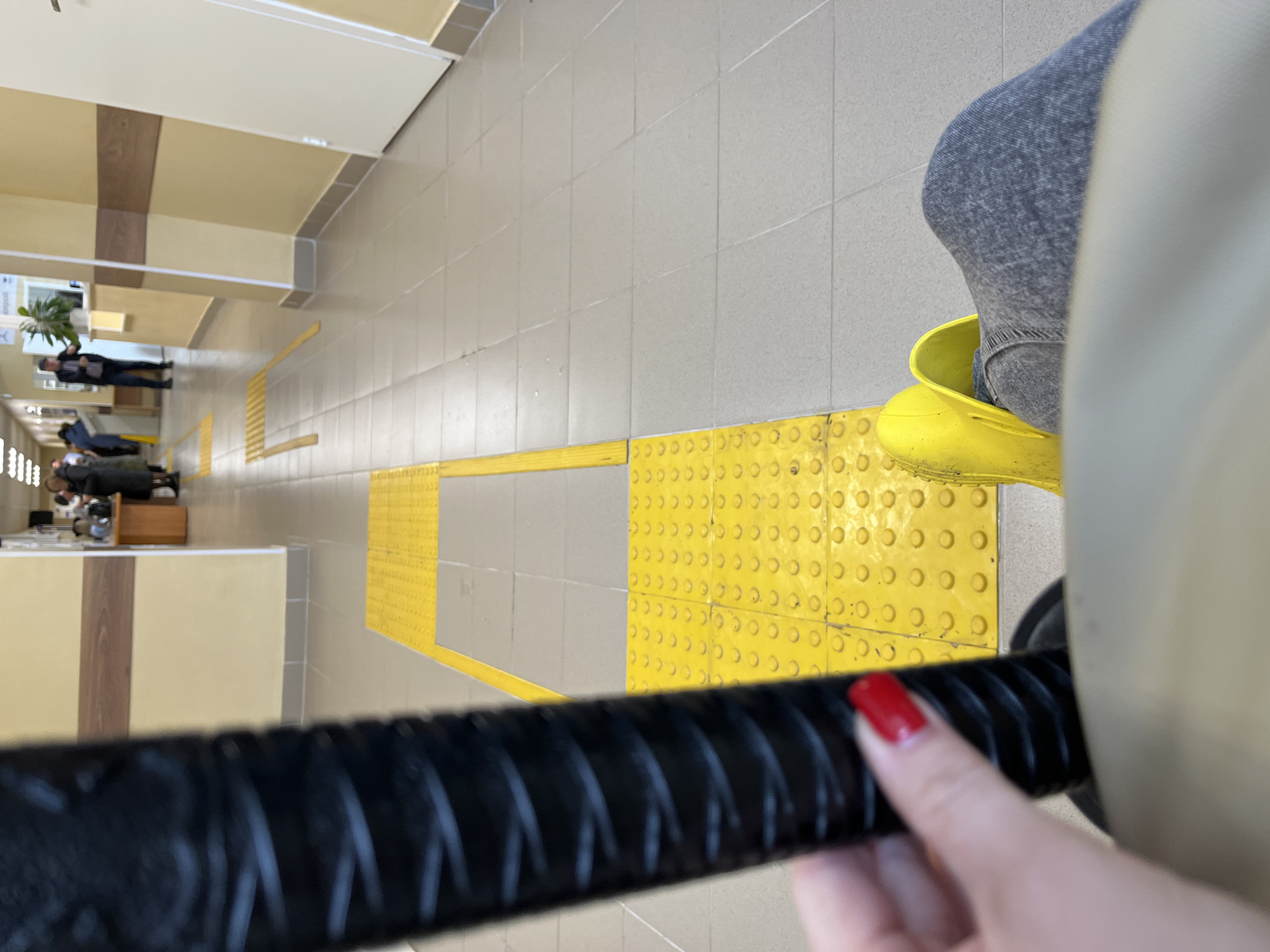 The image shows a hallway in what appears to be a public building, likely in Chaikovsky, Russia. The floor is tiled in grey and has yellow tactile paving strips. In the background, people are seen lying on benches. In the foreground, a person is wearing grey jeans and bright yellow Crocs-style shoes. A hand with red nail polish is holding a black rubber handle. The overall perspective suggests the photo was taken from a raised position, possibly from the top of a staircase or a raised platform.
