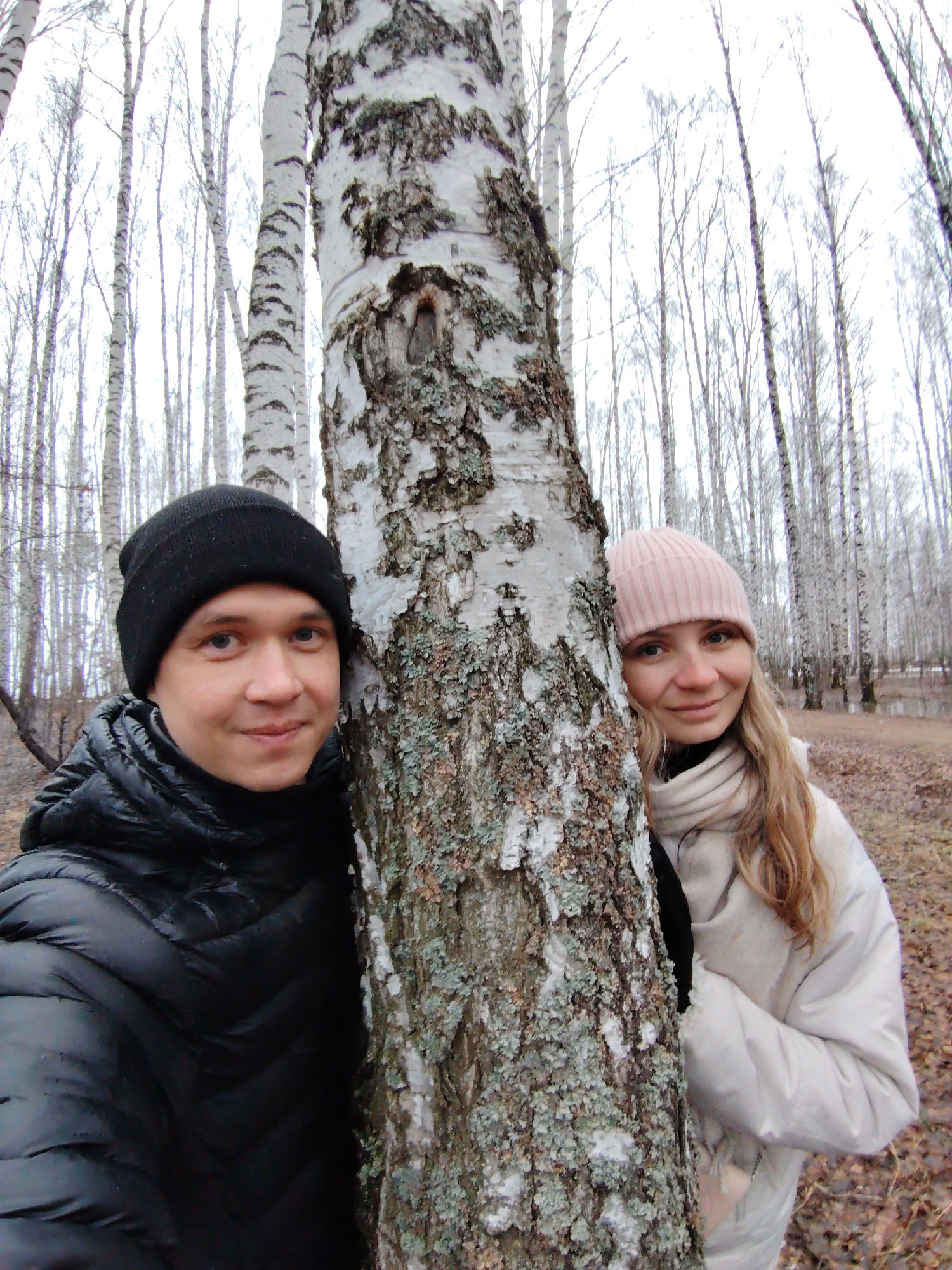 The image shows a man and woman posing behind a birch tree. The man is on the left side of the tree and is wearing a black winter hat and black puffer coat. The woman is on the right side of the tree and is wearing a pink beanie, a white scarf, and a white winter coat. The birch tree has a distinctive white bark with black markings. Behind the couple are several birch trees without leaves. The ground has dead leaves and there is a slight elevation in the background. The photo appears to be taken in a park or wooded area in Chaikovsky, Russia.