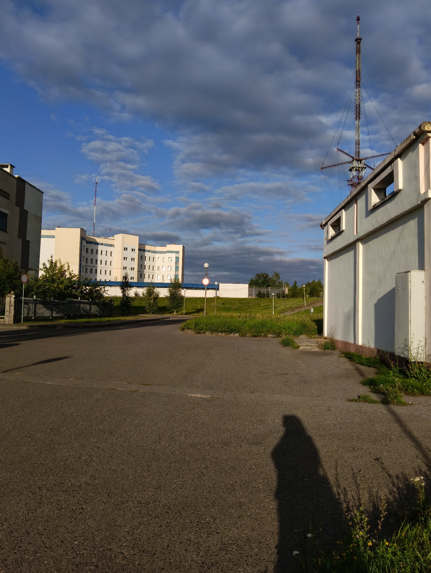 The image captures an outdoor scene in the city of Hrodna, Belarus, likely during the daytime under a partly cloudy sky. In the foreground, a dark asphalt surface is visible, with a distinct shadow of a person cast on it, indicating the presence of the photographer or another individual. To the right, a portion of a light-colored, rectangular building with pink accents along its top edge is prominent. This building has several small, rectangular openings near its roofline. Further into the background, a tall, lattice-like transmission tower stands against the sky. Beyond that, a modern, multi-story building with a cream-colored facade and numerous windows is seen. A black metal fence runs along the front of this building, behind which are scattered trees and green grass. The overall setting suggests an urban or semi-urban environment with a mix of residential or institutional buildings and infrastructure.