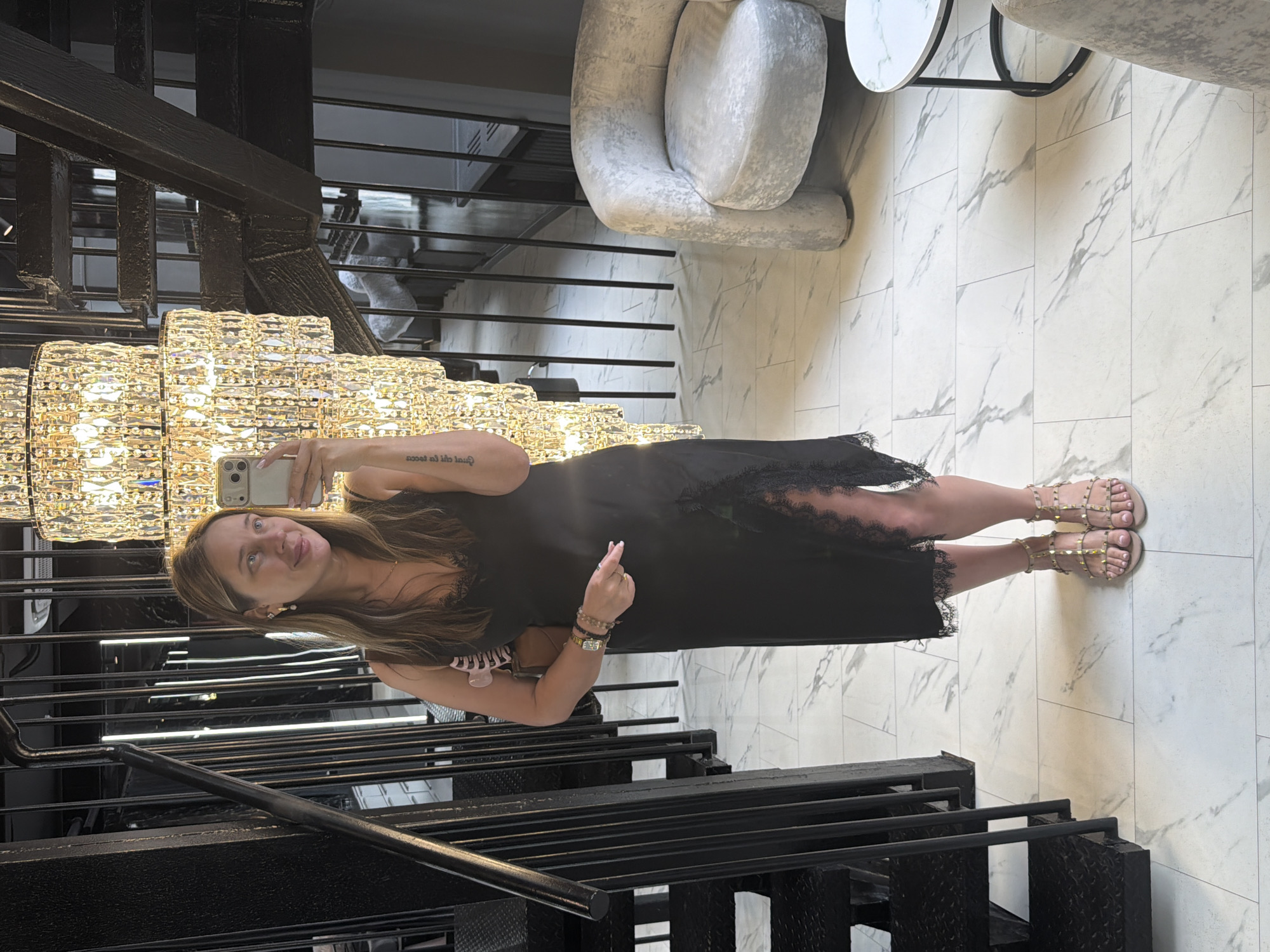 A woman is taking a mirror selfie in what appears to be a luxurious interior. She is wearing a black slip dress with lace trim and bejeweled sandals. She has long, light brown hair and is wearing earrings and bracelets. A handbag is slung over her shoulder. The background features a grand, multi-tiered crystal chandelier, a black metal staircase, and modern marble-patterned flooring. To the right, a plush grey armchair and a small round side table are visible. The overall impression is one of opulence and contemporary design. The provided location of Shcherbinka, Russia, is an external detail not discernible from the image itself.