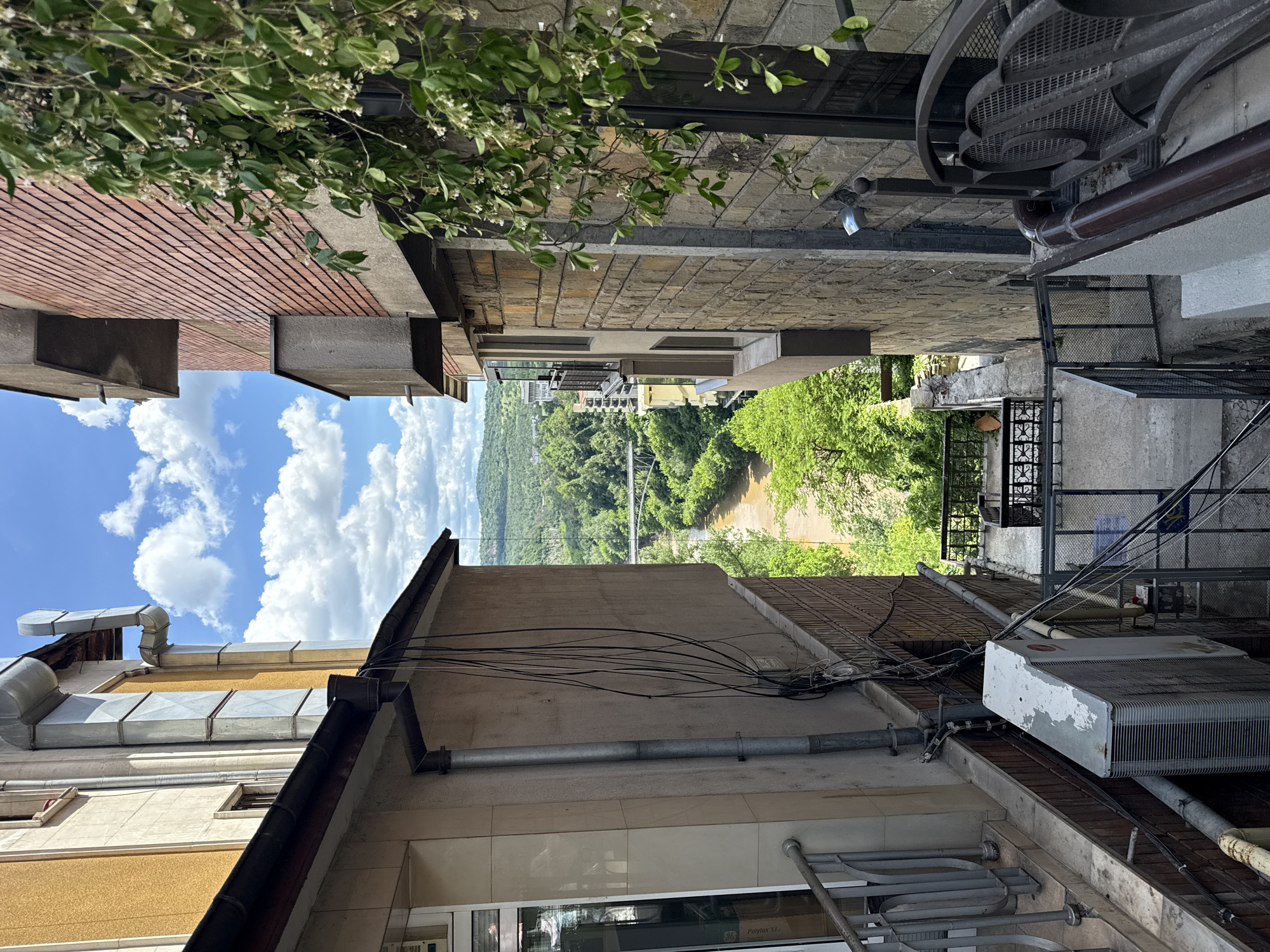 The image presents a view through a narrow passage between buildings, likely in the city of Veliko Tarnovo, Bulgaria. The angle suggests a high vantage point looking down and outwards, with elements of urban architecture framing a distant vista of a river valley and bridge.
On the left, there is a building with a brick facade and visible architectural elements such as gutters and ventilation ducts. The presence of cables and an air conditioning unit on the right-side building suggests a mix of older and more modern infrastructure. The sky is visible between the buildings, filled with fluffy white clouds.
In the distance, there is a river flowing through a lush valley, bordered by dense green trees. A bridge spans the river. The overall impression is of a built environment nestled within a natural landscape.