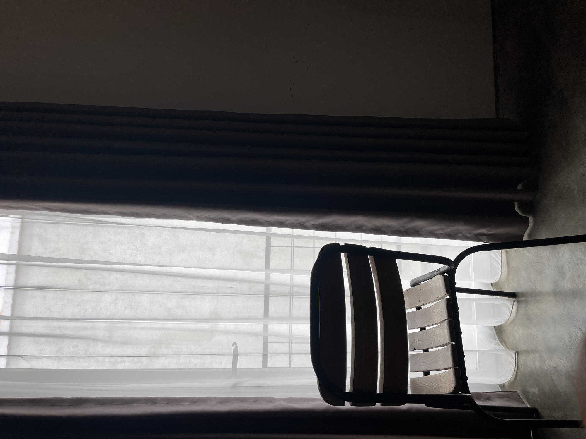 A dimly lit indoor scene shows a wooden chair with a black metal frame in front of a window covered with sheer white curtains and heavy brown drapes. The floor appears to be concrete. The location is suggested to be North Kuta, Indonesia, but there is no concrete visual evidence to confirm this. There are no people visible in the image. The chair is the primary object, situated in a way that suggests it is either placed near the window or the image is focused specifically on the chair. The curtains are drawn partially, filtering the light. The overall environment is dark.