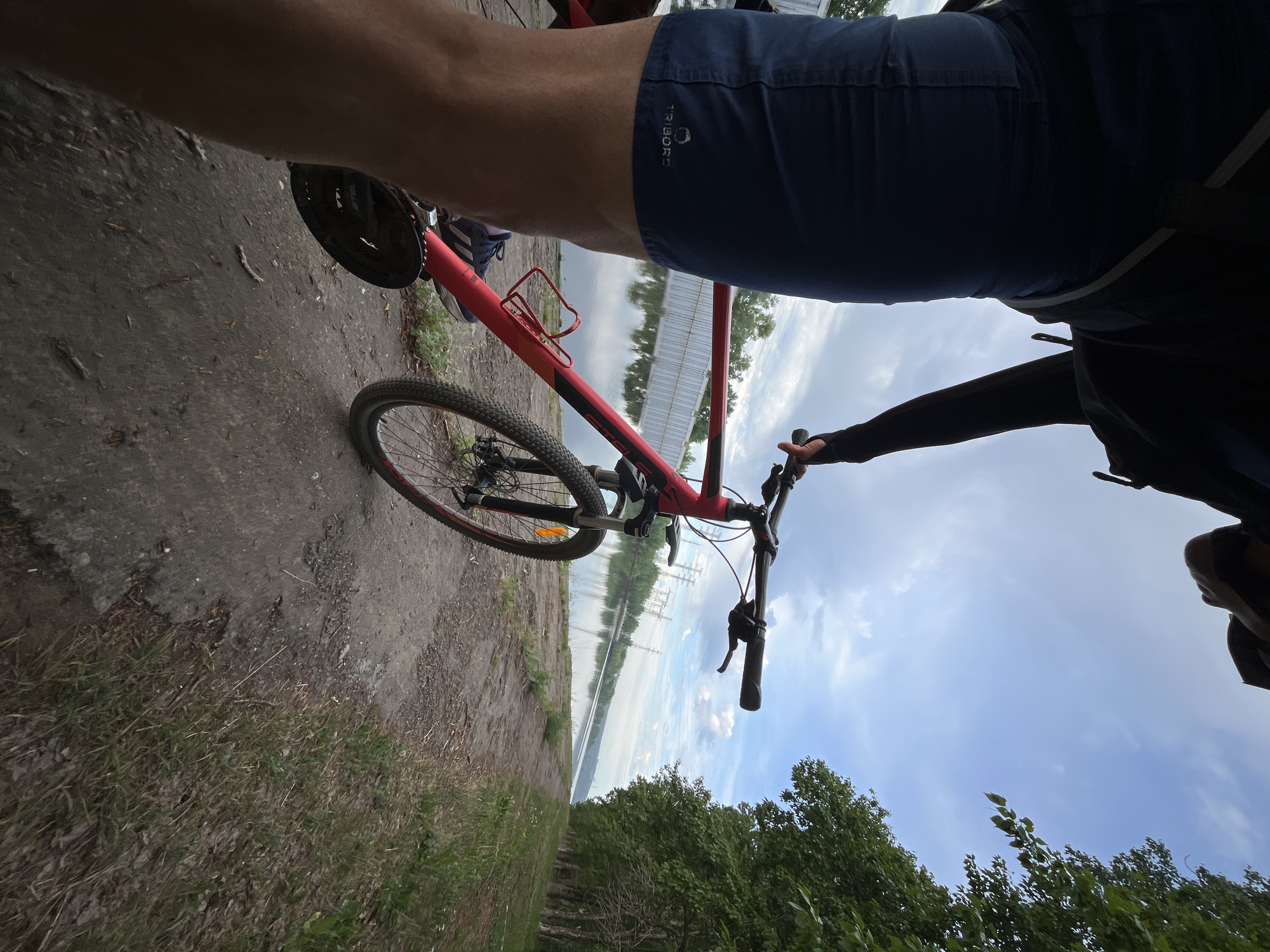 A low-angle, upside-down point-of-view shot shows a person on a red mountain bike. The person is wearing dark blue shorts with a white logo, and a dark long-sleeved shirt or jacket is also visible. The bike is on a dirt path next to green grass and trees. In the background, across what appears to be a body of water, there are utility poles and a fence. The sky is visible, with clouds. The location is Chaikovsky, Russia.