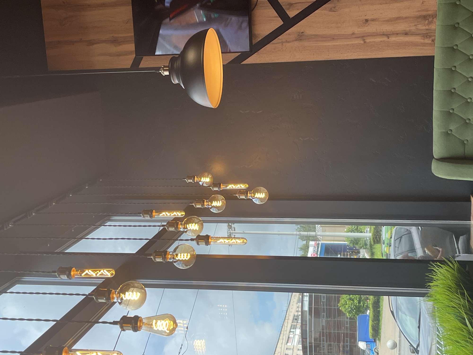 The image depicts an interior scene with large windows offering a view of an urban environment. A series of Edison-style light bulbs hang from the ceiling, illuminating the space with a warm glow. These bulbs vary in shape and size, some being round and others elongated tubes, all featuring visible filaments. A prominent black dome-shaped pendant light with a bright orange interior hangs on the right side of the frame. The walls are a dark gray, contrasted by a section of wooden paneling. Through the windows, a cityscape is visible, including buildings, trees, a blue truck, and cars. One of the cars, a dark SUV, has a person visible inside holding a package. The overall impression is of a modern establishment, possibly a cafe or restaurant, with a stylish industrial aesthetic. The presence of specific street signs and architectural styles visible through the window suggests the location is within the city of Izhevsk, Russia.