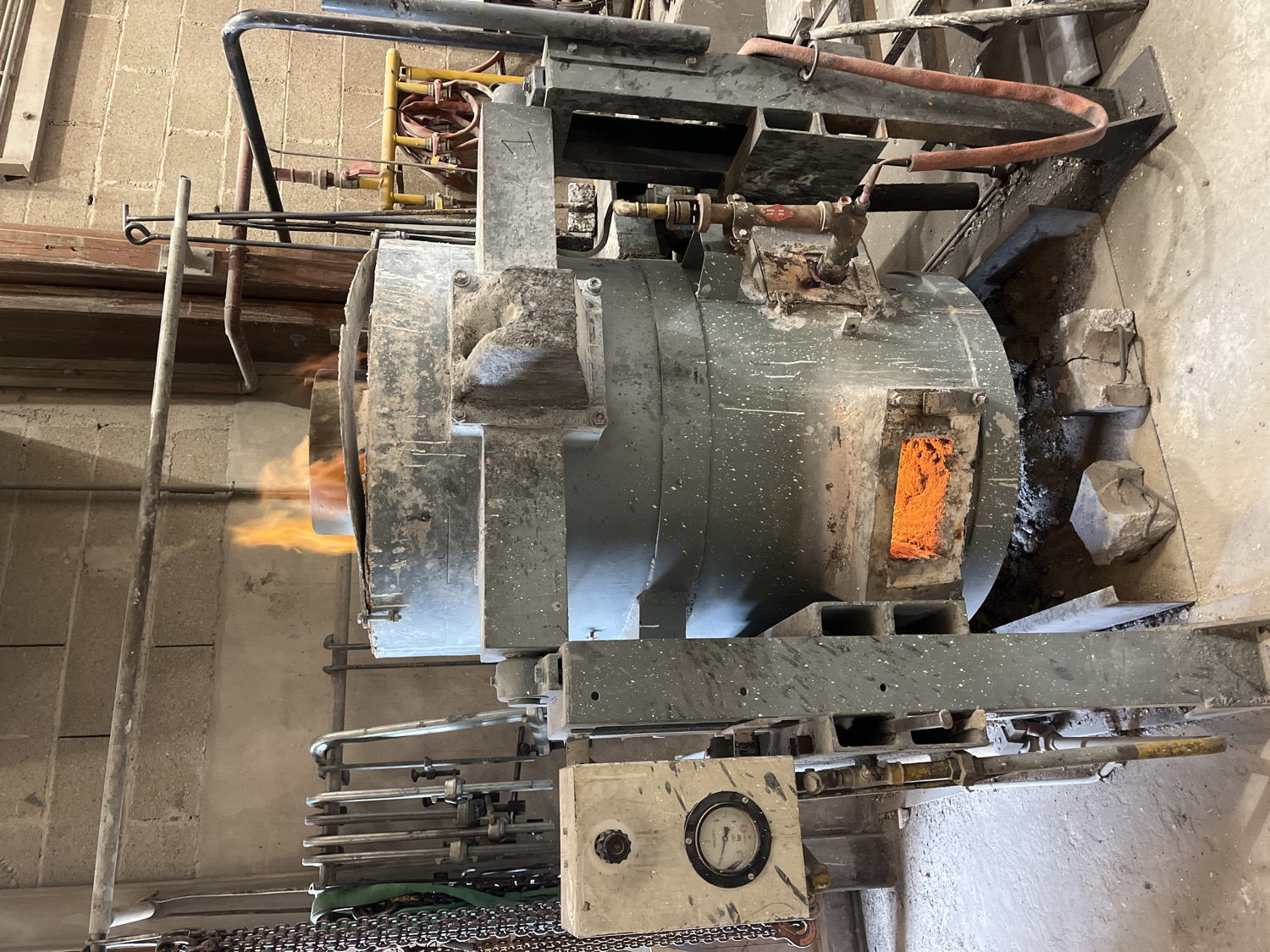 An industrial furnace or kiln, constructed primarily of sturdy grey metal, dominates the scene in an upright, portrait orientation. Flames, bright orange and yellow, vigorously emanate from an opening on the left side of the furnace, indicating active operation. On the opposite side of the furnace, a rectangular opening reveals an intense, glowing orange interior, suggesting the presence of molten material or extremely high temperatures within.
The furnace is integrated into a complex setup of pipes, hoses, and supporting metal structures, indicative of an industrial process. A thick, red-orange hose arches over the top of the furnace, while various smaller metal pipes and valves are visible throughout the machinery. Below the furnace, a small control box with a circular gauge is present, although specific readings or text on the gauge are not discernible. The ground around the furnace shows concrete blocks and debris, further emphasizing the industrial, working environment. In the background, a light-colored brick or concrete block wall is visible. The absence of people suggests a focus on the machinery itself. This scene depicts a high-temperature process, likely in a workshop or small factory, in Saint-Rémy-lès-Chevreuse, France.