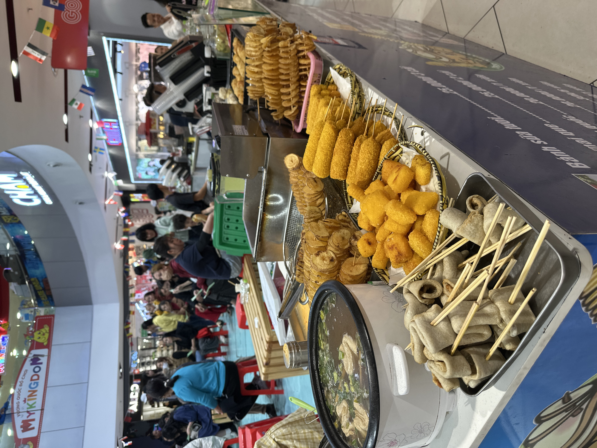 This is a photograph taken at a street food stall in Da Lat, Vietnam. The scene is bustling with people, suggesting it is a popular spot, possibly in a market or shopping area, given the presence of a "MYKINGDOM" sign, which indicates a toy store, hinting at a family-friendly environment.
The food stall displays a variety of popular street foods. Prominently featured are spiral-cut fried potatoes on skewers, corn dogs coated in a golden batter, and what appear to be fish cakes or similar items served on skewers in a metal tray. A pot of stew with visible ingredients like meat and green onions sits at the front of the stall. The food is arranged attractively, some in baskets and trays, designed to entice customers.
Several people are visible in the background, some browsing the food, others potentially shopping or passing through. The lighting suggests it could be daytime or an indoor setting with artificial lights, as is common in Vietnamese markets. The overall atmosphere seems lively and energetic, indicative of a busy commercial area. The image also shows several small national flags strung up, suggesting a possible international theme or location catering to diverse visitors. The text visible includes "JONG QUỐC ĐỒ CHƠI" and "MYKINGDOM", along with some Vietnamese text that appears to be menu items or descriptions like "Banh sửa to" and "mai quen".