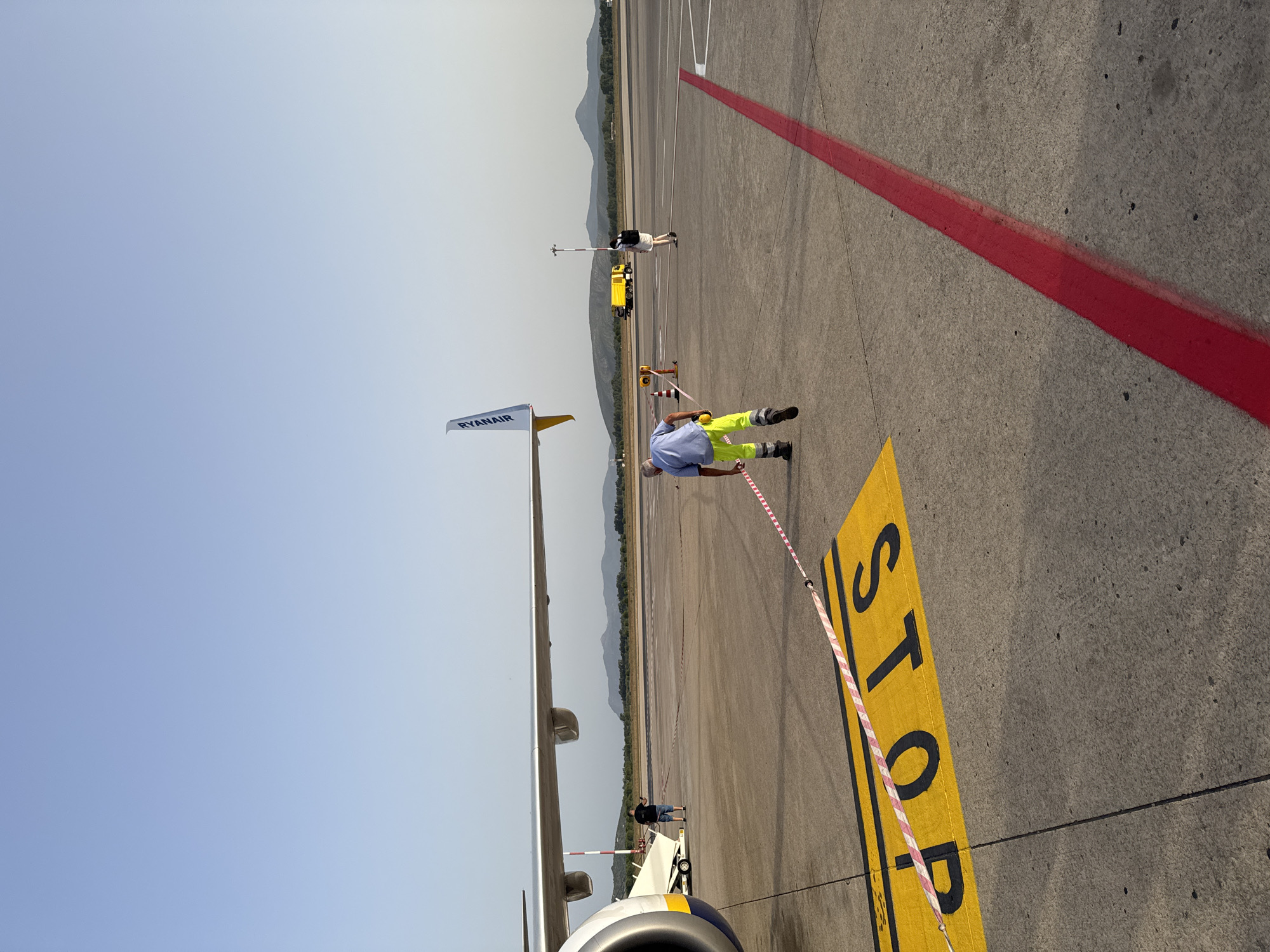 The image depicts an airport tarmac at Alghero, Italy. In the foreground, a large yellow "STOP" marking is visible on the asphalt, partially obscured by a red and white striped barrier tape. To the left, a portion of a Ryanair airplane, including its engine and wing, is present. A person wearing high-visibility green trousers and a blue shirt is walking away from the camera, pulling the barrier tape. Further in the background, other individuals, a yellow service vehicle, and hills are visible under a clear blue sky. The scene suggests an operational environment on the tarmac with ongoing activities.