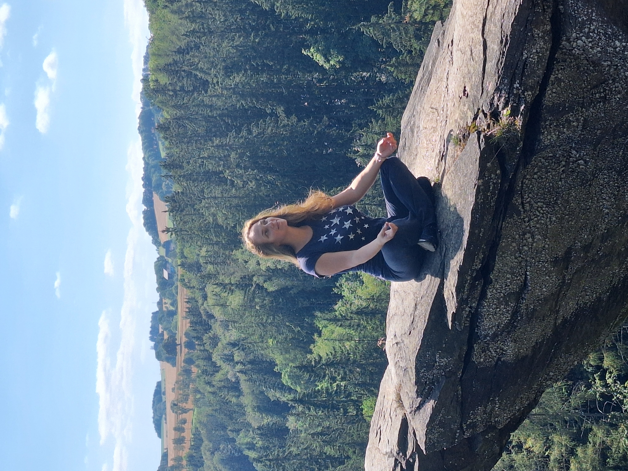 A woman is meditating on a rock formation with a forest in the background, near Wolkenstein, Germany. The woman is sitting in a lotus position, wearing a dark blue t-shirt with white stars and dark pants. She has long, wavy blonde hair. The rock formation is large and appears to be part of a cliff edge. The background consists of a dense forest with evergreen trees and a hilly landscape beyond. The sky is blue with scattered white clouds. The image seems to be taken from a slight low angle, emphasizing the height of the rock formation and the expanse of the landscape.