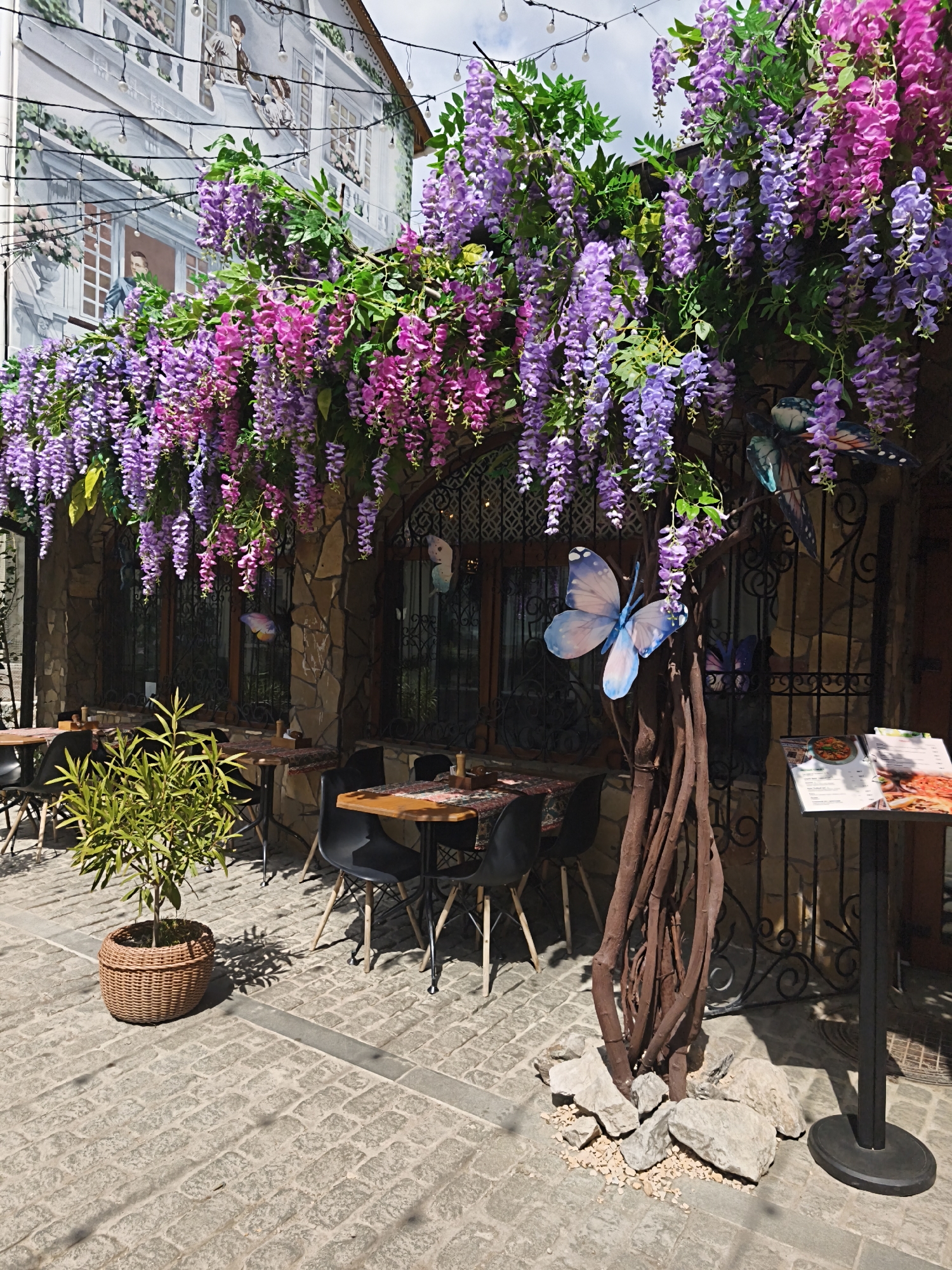 The image depicts an outdoor seating area in what appears to be a European-style restaurant or cafe in Алупкинский городской совет, Ukraine. The foreground shows a paved stone surface leading to several tables and chairs, suggesting a dining space. A potted plant sits to the left, and a decorative tree-like structure with artificial purple and pink wisteria hangs above the seating area. Ornamental butterflies are attached to the tree. The building behind the seating area has a stone facade with windows and wrought iron details. A menu stand is visible near the right edge of the frame. A mural is visible on an adjacent building depicting figures on balconies. The scene is bathed in sunlight, indicating a daytime setting.