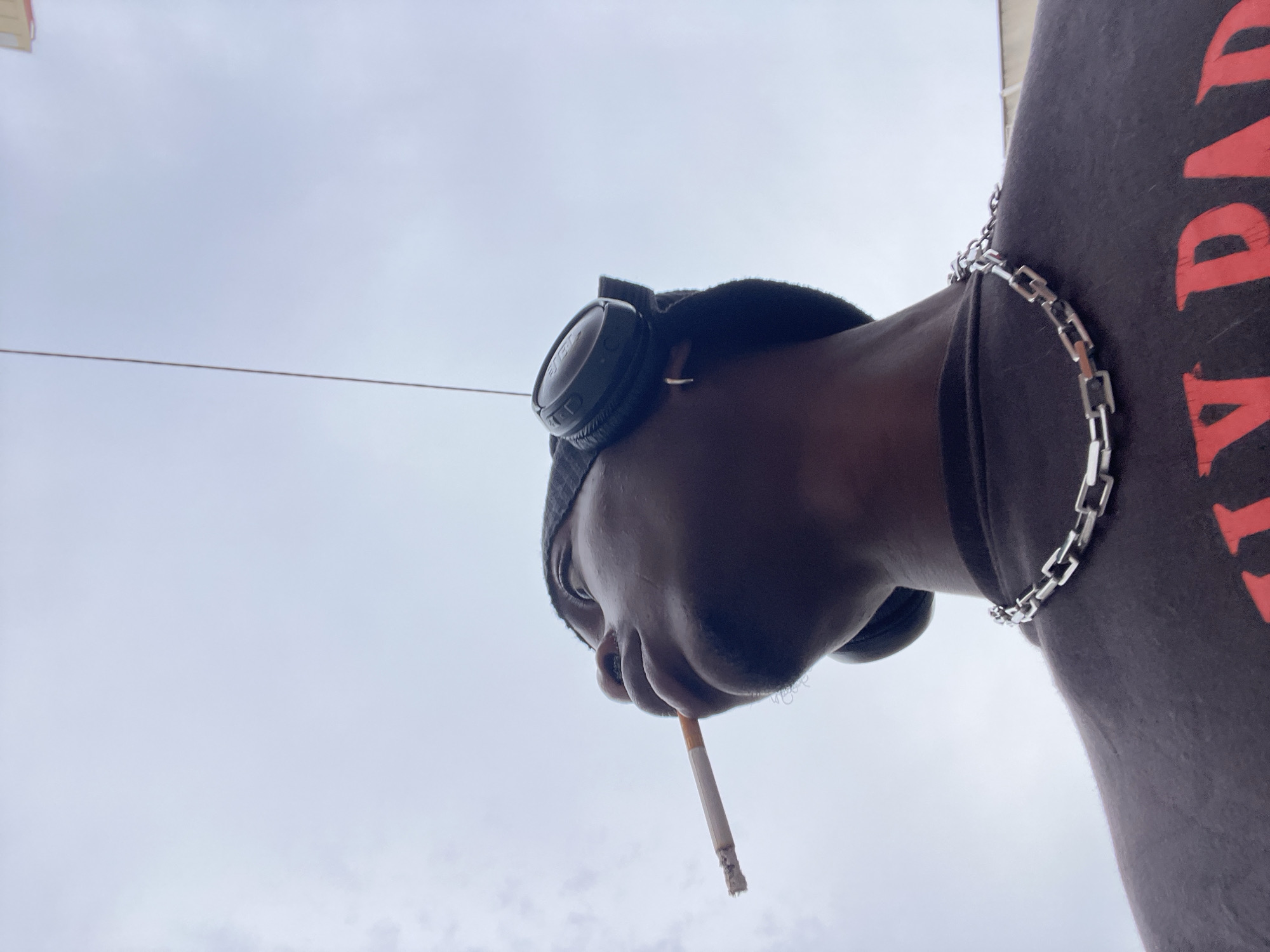 A low-angle shot captures a person smoking a cigarette against a cloudy sky. The person is wearing headphones, a beanie, a silver chain necklace, and a dark-colored shirt with a red design on the right side of the frame. The person's face is angled downwards, cigarette in mouth. The location is Barcelona, Spain.