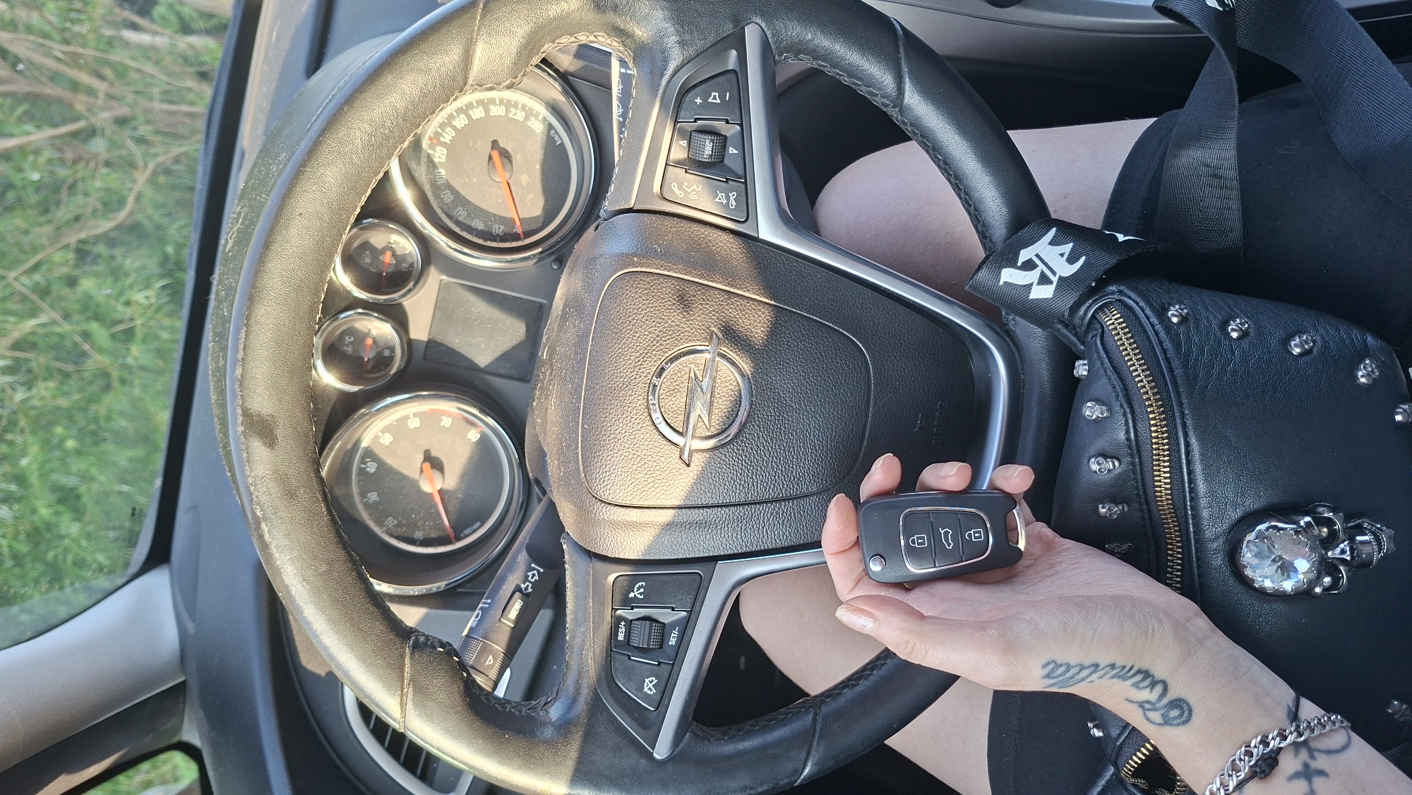 A person is sitting in the driver's seat of a car, holding a car key in their left hand. The car appears to be an Opel, with the logo visible on the steering wheel. The dashboard and steering wheel are in the foreground, with the natural environment visible through the windshield. The person is wearing a black fanny pack decorated with skull studs. A tattoo of the name "Kamilla" is visible on their forearm. The image is likely taken in Myadzyel, Belarus, as indicated by the location.