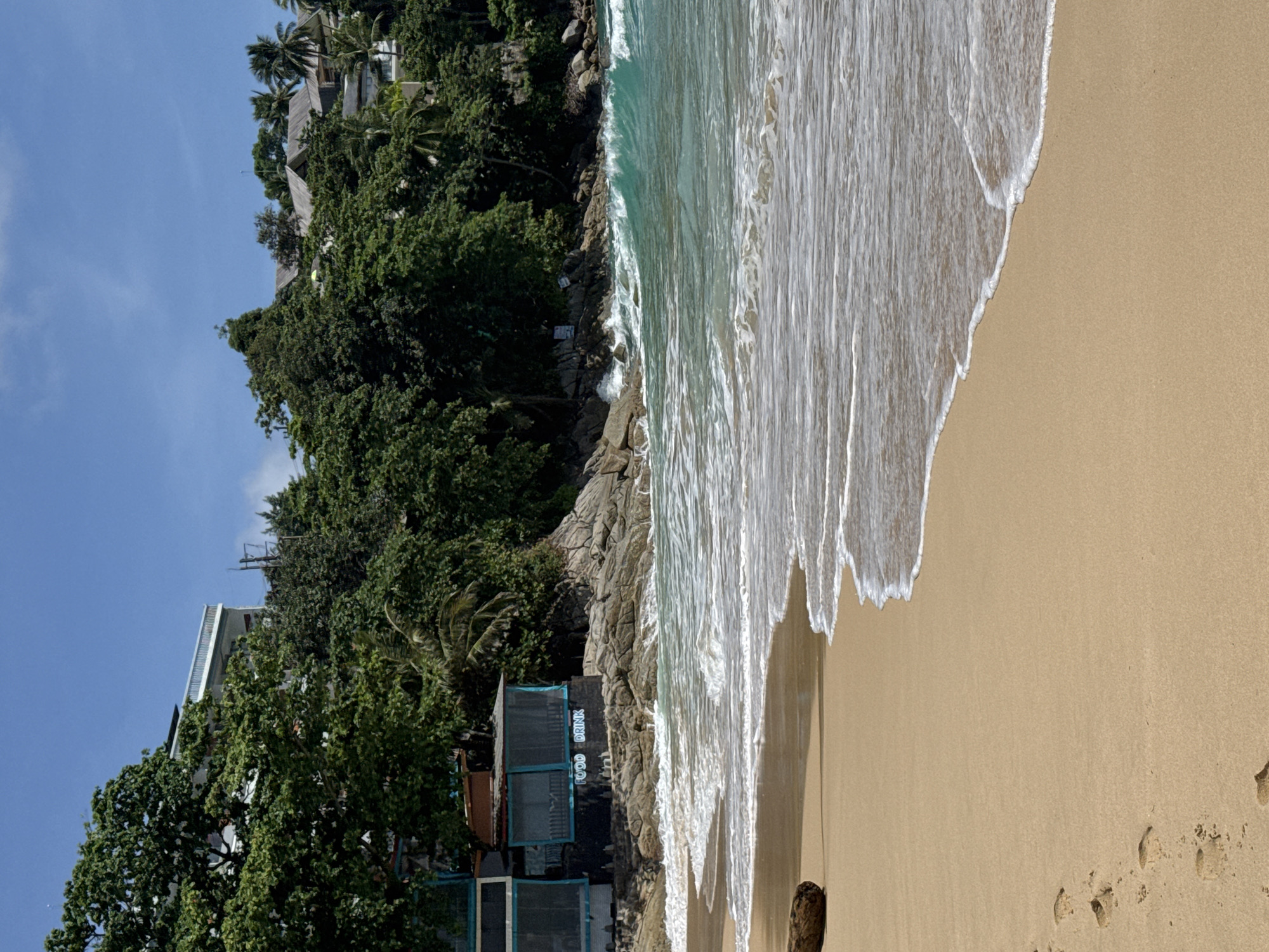 The image depicts a beach scene in Choeng Thale, Thailand, under a clear blue sky with scattered clouds. The foreground is dominated by a sandy beach with several footprints leading away from the water. A piece of driftwood rests on the left side of the sand. The ocean is visible with waves breaking on the shore, creating white foam. The water is a turquoise color. In the background, a rocky area leads up to a lush green hillside covered in dense trees. A building with a white railing and several smaller buildings with wooden roofs are partially visible amongst the foliage. A small structure with a black facade and blue trim, labeled "FOOD DRINK", is situated near the rocky shoreline.