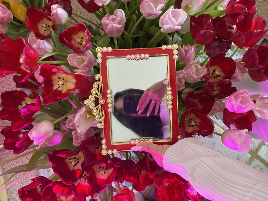 The scene is an indoor close-up located in Chaikovsky, Russia. A person's left hand, adorned with pink-tinted manicured fingernails and clad in a white, vertically pleated sleeve, holds a small, ornate rectangular mirror with a red frame. The frame features gold accents and faux pearls along its edges and a more elaborate, crown-like gold and pearl design at the top center. Reflected in the mirror is the upper torso and head of a person with long dark hair, wearing a white garment. The person's right hand, displaying a ring on the ring finger, holds a dark-colored smartphone, indicating a selfie being taken via the mirror. The background is filled with a dense arrangement of natural tulips in various stages of bloom, predominantly deep red and light pink, with visible green stems and leaves. A reflective surface with a faint geometric pattern is partially visible in the lower left portion of the image. The environment is brightly lit with a distinct pinkish-purple color cast.