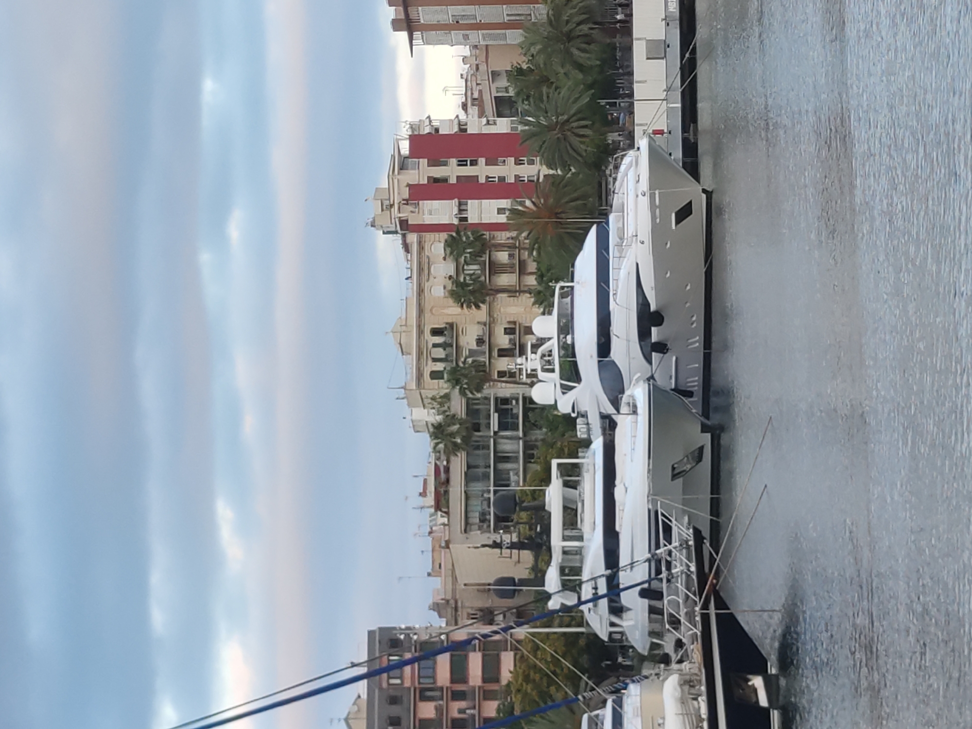 The image captures a view of yachts docked in a harbor, with buildings visible in the background. The sky is overcast with soft clouds, suggesting it might be late afternoon or early morning. The water in the harbor is calm, reflecting the sky and the boats. In the foreground, two large white yachts are prominently featured, appearing to be luxury vessels. One yacht is positioned slightly in front of the other. A dark-hulled boat is partially visible on the left side of the frame. The buildings in the background are a mix of modern and older architecture, some with balconies and windows. Palm trees are scattered among the buildings, indicating a warm climate. The overall scene is one of a serene, well-maintained marina. This scene is likely located in the Port Olímpic area of Barcelona, Spain, which is known for its upscale marinas and modern waterfront development, often hosting luxury yachts and surrounded by contemporary architecture.