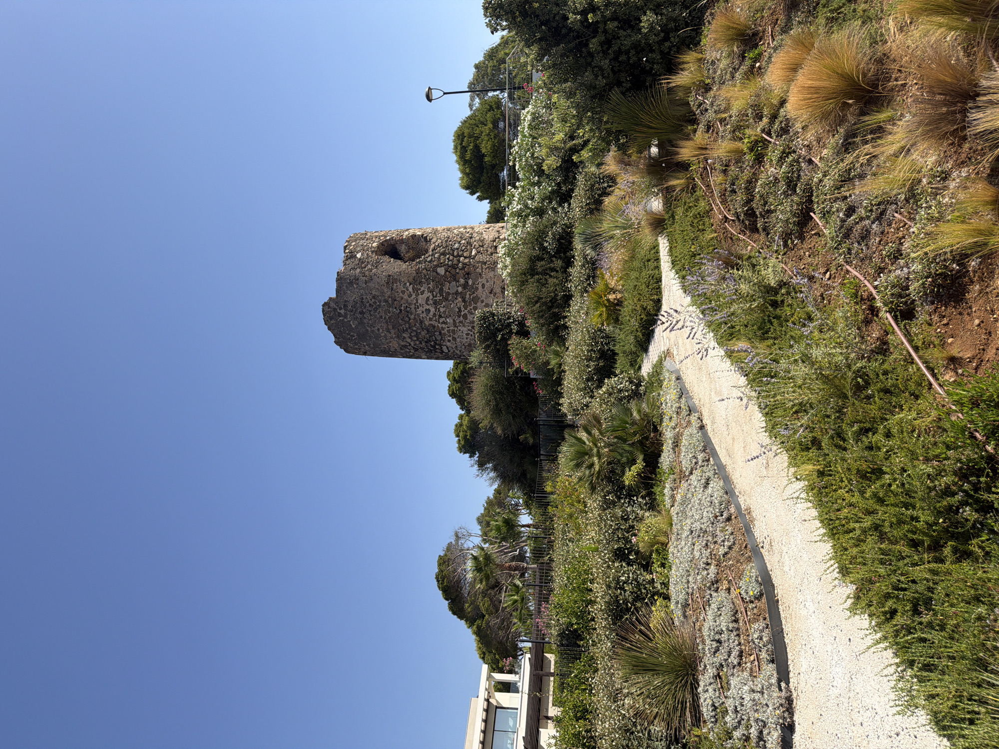The image depicts a historical stone tower, likely a watchtower, situated atop a hill in Estepona, Spain. The tower is weathered and circular, with a large opening visible on its side. It is surrounded by lush, Mediterranean vegetation, including shrubs, grasses, and some palm trees. A paved pathway winds through the greenery, leading towards the tower. In the background, a modern building with large windows and a dark pergola is visible, suggesting a residential or resort area. A street lamp stands near the tower. The sky is a clear, bright blue.