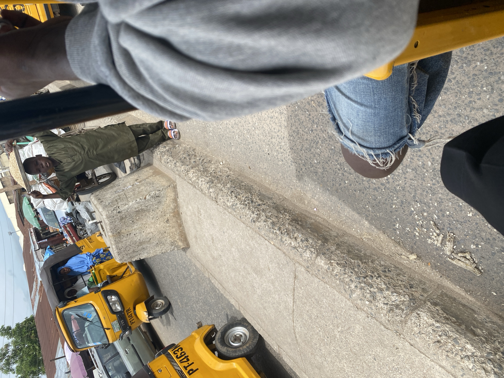 The image depicts a street scene in Jalingo, Nigeria, likely in a market area. In the foreground, a person wearing ripped blue jeans and a grey hoodie is seated, with a yellow vehicle, possibly a tricycle taxi, visible. To the left, another yellow tricycle taxi is parked, with the license plate "PT-463N" visible. In the middle ground, a young boy dressed in a green outfit stands near a cart laden with goods, possibly food items. Behind him, a yellow tricycle taxi is occupied by two women wearing traditional headscarves. The background reveals market stalls with corrugated iron roofs and a dusty, urban environment under an overcast sky. The ground is paved with asphalt and concrete, with a noticeable curb separating the road from the sidewalk.