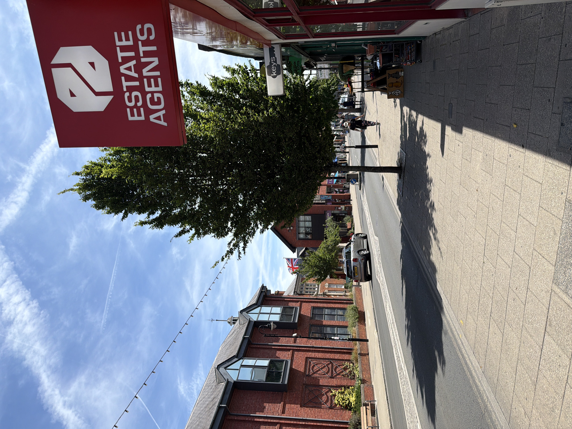The image depicts a street scene in the United Kingdom on a sunny day. A red sign for "ESTATE AGENTS" with a white logo is prominently displayed on the right side of the frame. A mature tree with lush green foliage stands next to the sign, casting a shadow on the sidewalk. The sidewalk is paved with rectangular stones, and on the left side of the street, a red brick building with large windows is visible. Further down the street, a silver car is driving away from the camera, and a Union Jack flag is flying from a building. Several people are walking on the sidewalk, and market stalls are set up on the right side of the street. String lights are hung across the street, suggesting a festive atmosphere or a commercial area. The sky is blue with scattered white clouds and contrails from an aircraft. The overall impression is that of a typical urban street in a town or city in the UK.