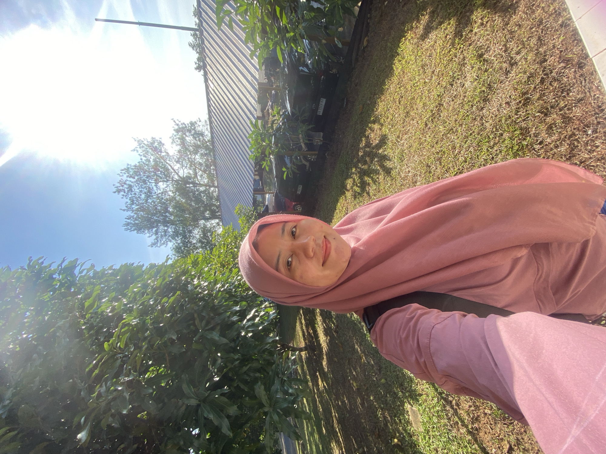 A young Indonesian woman wearing a pink hijab and a matching pink shirt is taking a selfie outdoors. She is smiling and looking at the camera. Behind her, there are trees with green leaves, a grassy area, and a building with a corrugated metal roof. Several cars are parked under the roof of the building. The sky is blue and the sun is shining brightly, casting shadows on the ground. The location is likely a public space in Banjarbaru, Indonesia, given the context of the images.