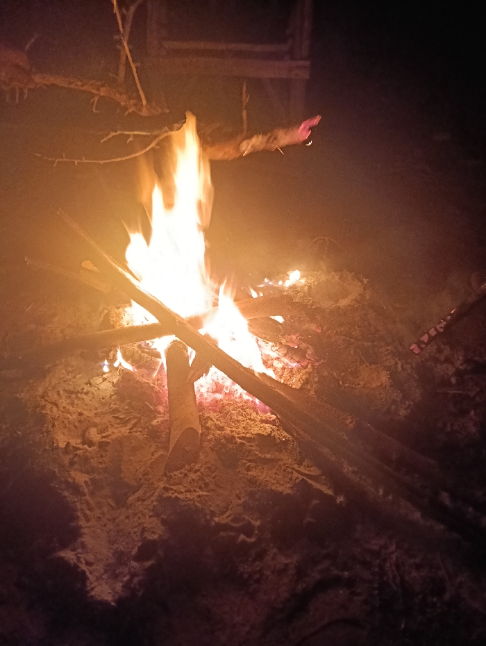 In Kyamatu, Kenya, a nighttime scene depicts a large, burning bonfire. The fire occupies the central and lower portions of the frame, emitting bright orange and yellow flames from a pile of logs and branches. The immediate ground around the fire appears to be disturbed soil or sand, illuminated by the fire's intense glow. In the upper-left quadrant, a human arm, with a visible hand and wrist, extends horizontally towards the flames from off-camera. This arm appears to be light-skinned and is slightly blurred, possibly due to motion or heat haze, indicating an interaction with the fire. The background is predominantly dark, with indistinct, shadowy forms suggesting a rudimentary wooden structure or large objects beyond the fire's reach, consistent with an outdoor, unlit environment.