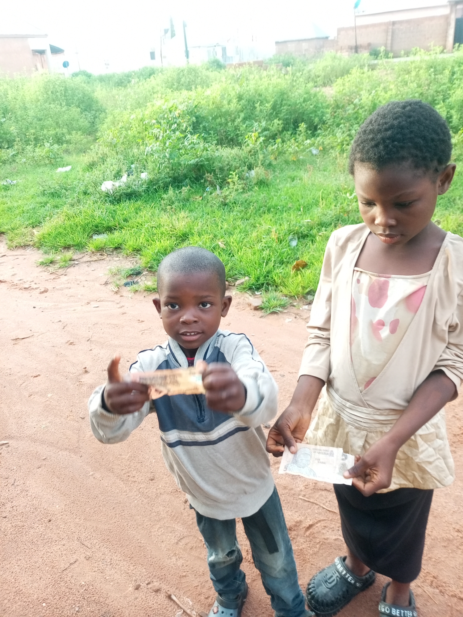 In Tempure, Nigeria, two children stand on a dirt path with a grassy embankment behind them. The child on the left, a young boy, wears a light grey sweatshirt with blue and black stripes and ripped blue jeans. He holds a crumpled banknote in his right hand, gesturing with it. The child on the right, a young girl, wears a beige dress over a patterned undergarment and a black skirt. She holds a similar banknote in her left hand. Both children appear to be looking at something off-camera. The environment is outdoors with natural light.