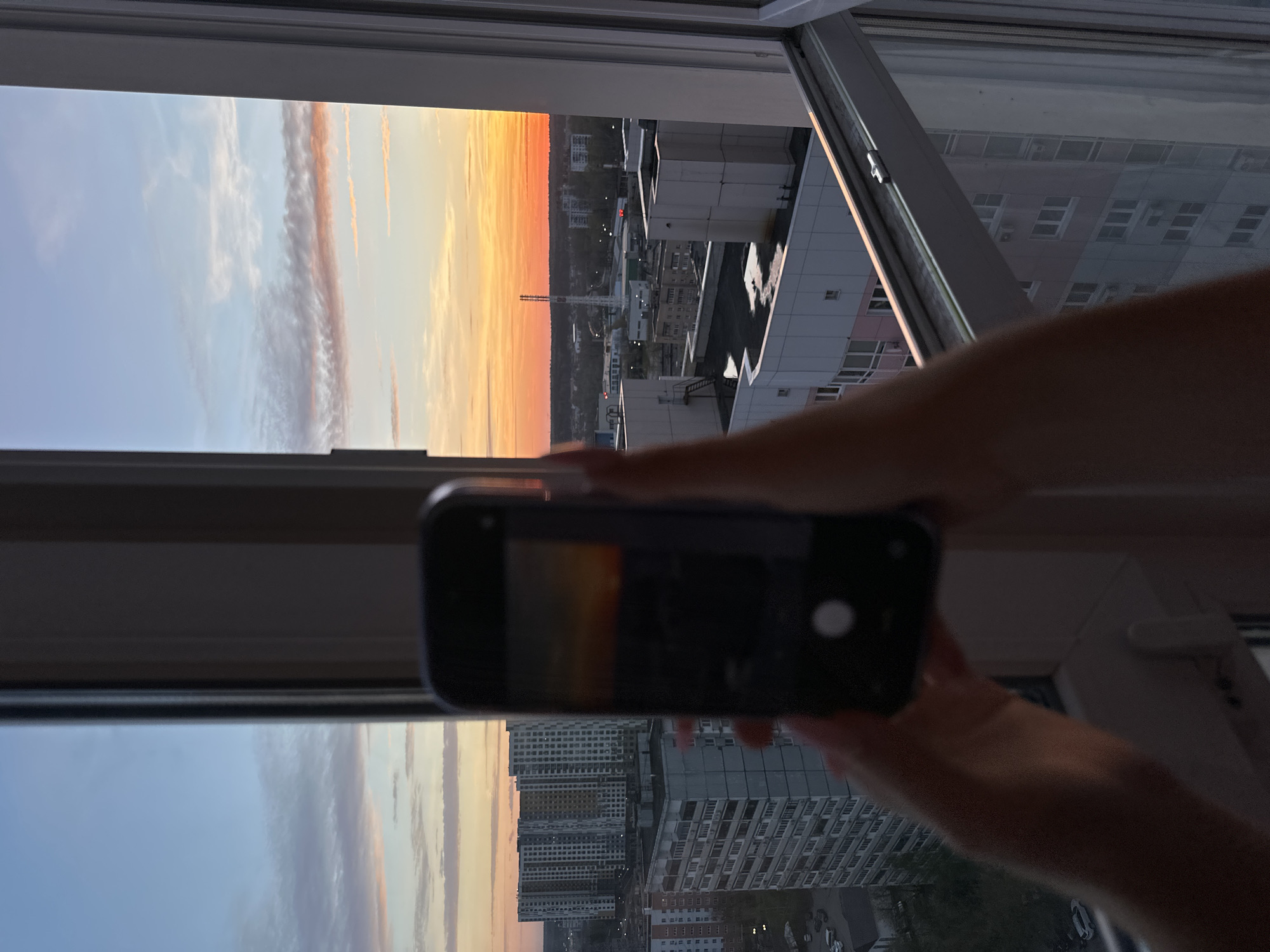 The image shows a sunset viewed through a window. An individual is taking a photo of the sunset with their smartphone. The scene outside the window includes residential buildings and a smokestack silhouetted against the orange sky. The photo is taken from an apartment in Moscow, Russia.