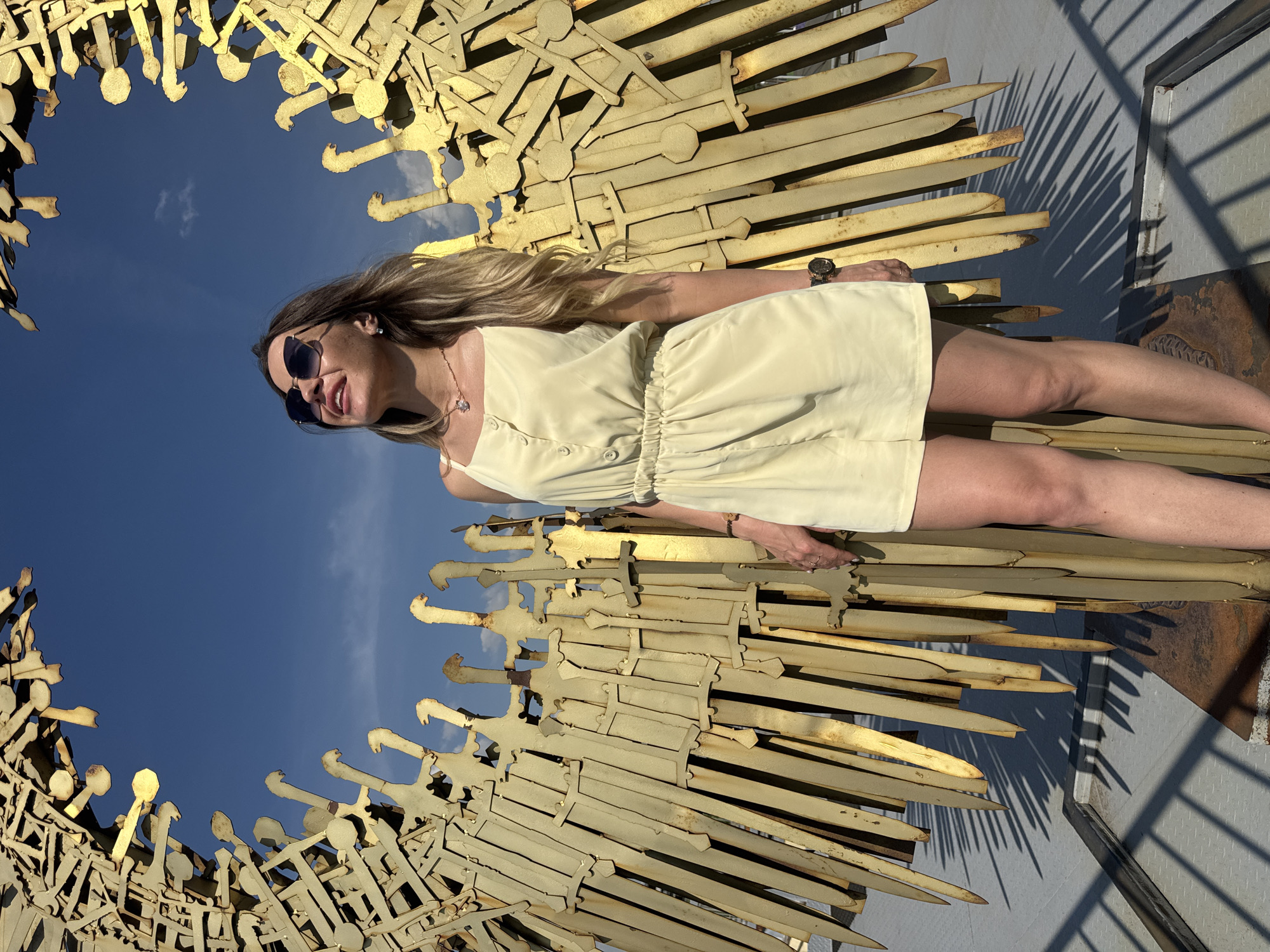 A woman stands in front of a large, golden sculpture made of what appears to be swords, which resembles wings. The sculpture is located outdoors, with a bright blue sky and some clouds visible. The woman is wearing sunglasses, a light-colored sleeveless romper, and a necklace. Her hair is long and blonde. The sculpture casts sharp shadows on the ground, which appears to be a paved surface. The overall impression is of a sunny day in an urban setting, possibly a park or public square in Saint Petersburg, Russia, given the presence of such monumental art.