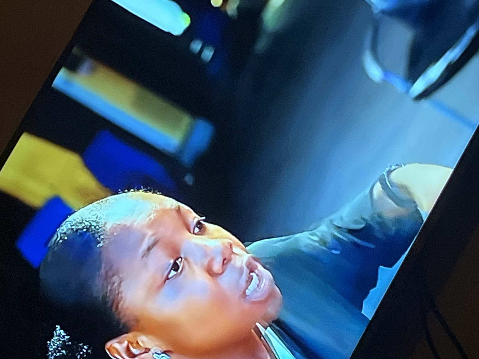 The image shows a close-up of a Black woman from the chest up, angled from her left side. Her hair is pulled back with some gelled edges. She is wearing a light blue earring in her visible left ear. Her mouth is open, and her teeth are visible, suggesting she is speaking or reacting to something. She is wearing a dark teal garment. The background is blurred with bright, abstract shapes of blue, yellow, and possibly green, indicative of a brightly lit environment with colorful elements. The lower portion of the image shows a dark surface, possibly a table or the edge of a screen, and a blurry dark object with blue and white accents that could be part of furniture or another screen. There is no discernible information within the image that would indicate a specific location in the city of Gura Top, Nigeria.