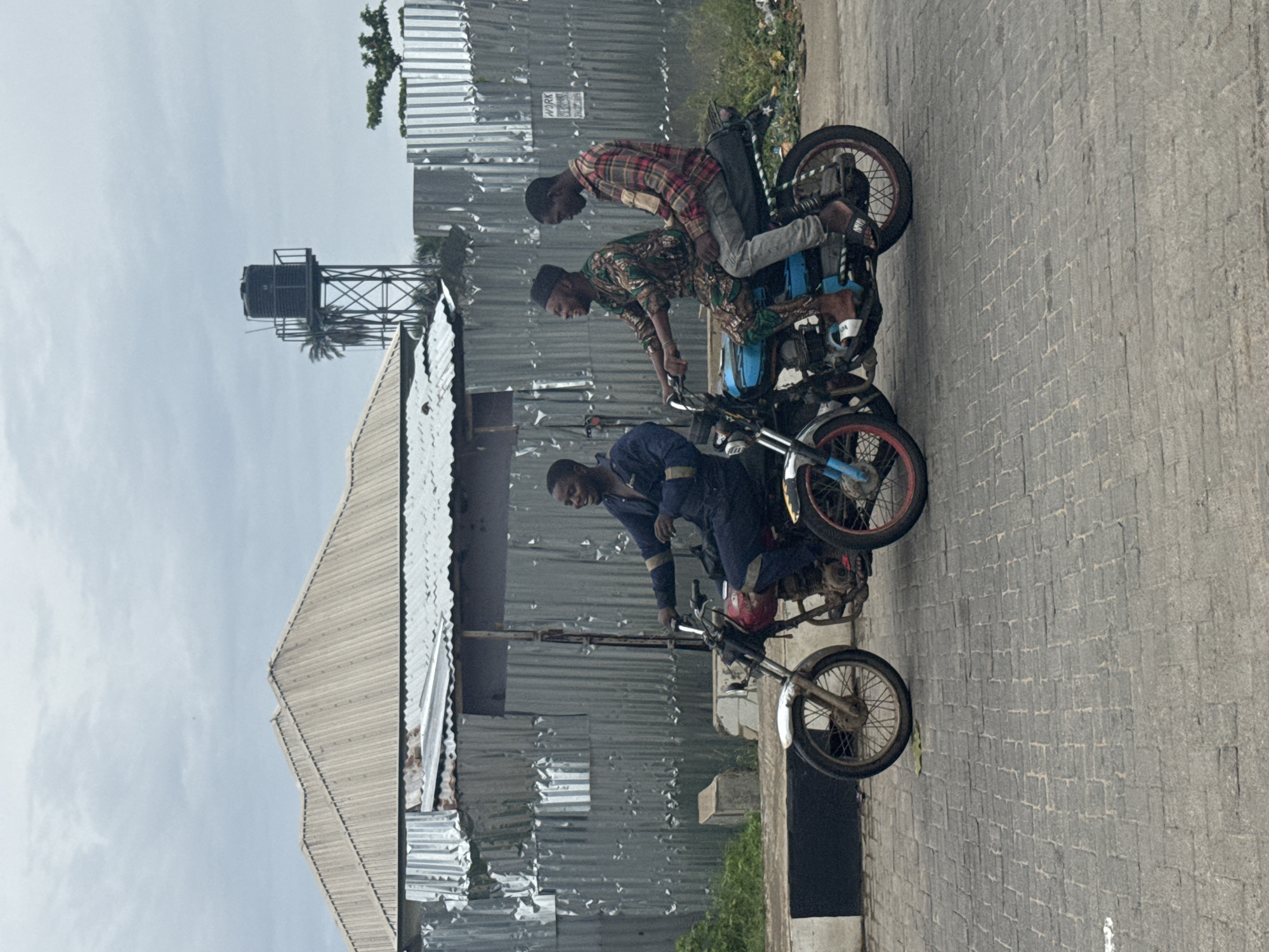 Three men are riding two motorcycles in Badagry, Nigeria. The first motorcycle has one rider wearing dark blue overalls. The second motorcycle has two men on it. The rider is wearing an ornate brown and green shirt, while the passenger is wearing a red plaid shirt and light grey pants. There is a corrugated metal building, a water tower, and trash in the background. The street is paved with bricks.