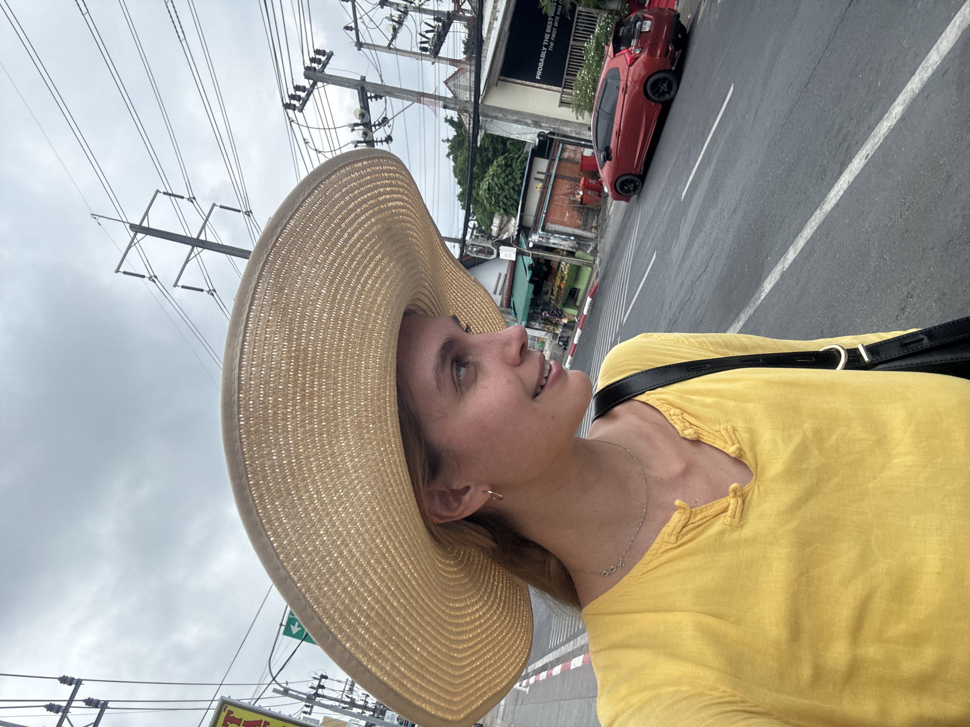 The image presents a woman standing on the side of a road. She is wearing a large-brimmed straw hat and a yellow shirt. She has a shoulder bag with a black strap. In the background, there is a red car parked on the side of the road, buildings, power lines and utility poles, and a cloudy sky. The scene is located in Patong, Thailand, as inferred from contextual clues related to local architecture and street layouts.