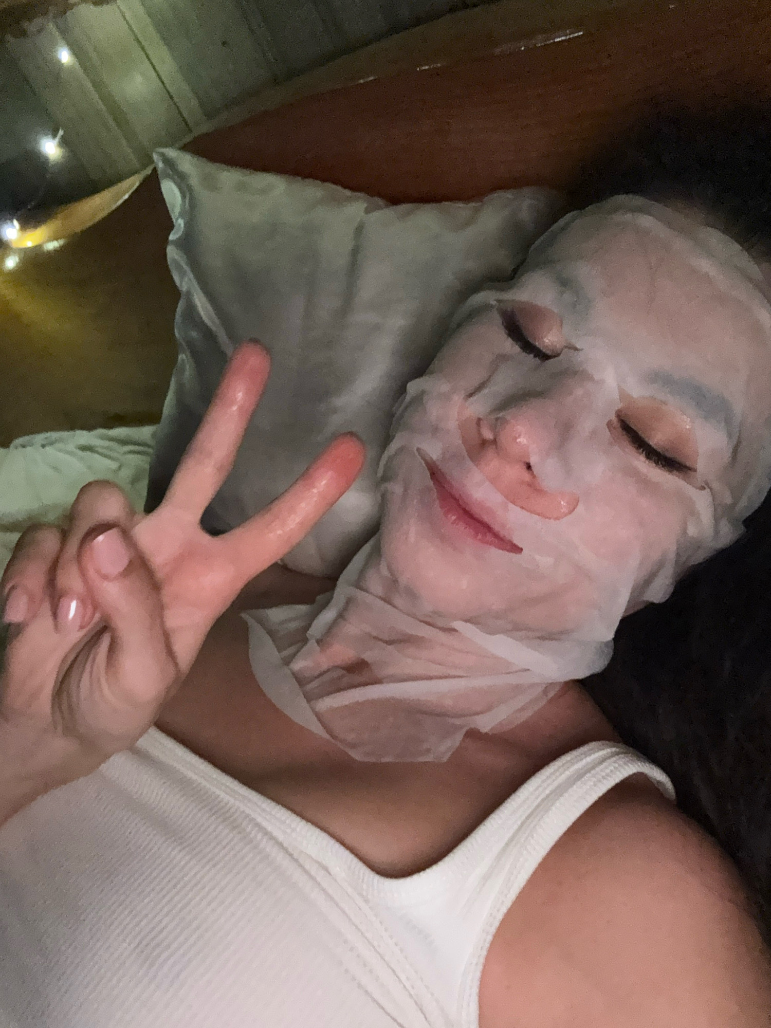 A woman is lying on her back, wearing a sheet face mask and a white ribbed tank top. She is holding up two fingers in a peace sign with her left hand. The background shows a pillow, a wooden ceiling with fairy lights, and a dark wall. The setting appears to be a bedroom. There are no identifiable landmarks of Hiệp Thạnh, Vietnam.