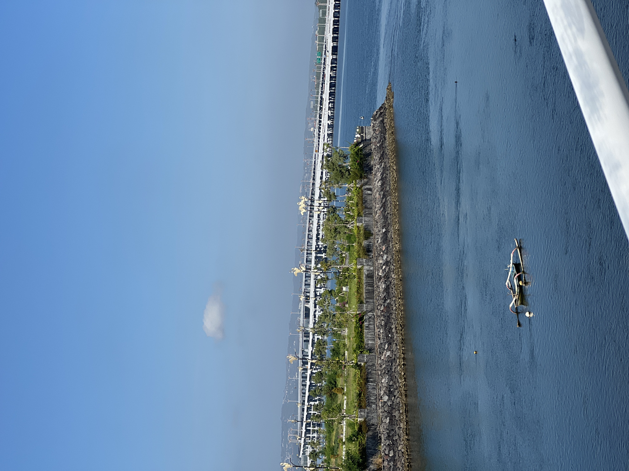 The image presents an elevated view, possibly from a bridge or tall structure, showing a section of coast adjacent to Denpasar, Indonesia. A narrow strip of land, acting as a breakwater or artificially constructed shoreline, runs horizontally across the image. This strip features rock stabilization, interspersed with vegetation, trees, and structures that suggest a park or planned development. A series of uniform, white structures with ornate lampposts line the inner edge of this strip, indicating a pathway or road.
Beyond this strip of land, the image reveals a large body of water, likely the ocean or a bay. A small boat or watercraft is visible in the water. Farther in the distance, a bridge or causeway stretches into the background, suggesting infrastructure for transportation connecting the area.
The sky is a clear blue with minimal cloud cover. The overall impression is a mix of coastal development and natural elements, with transportation infrastructure nearby.