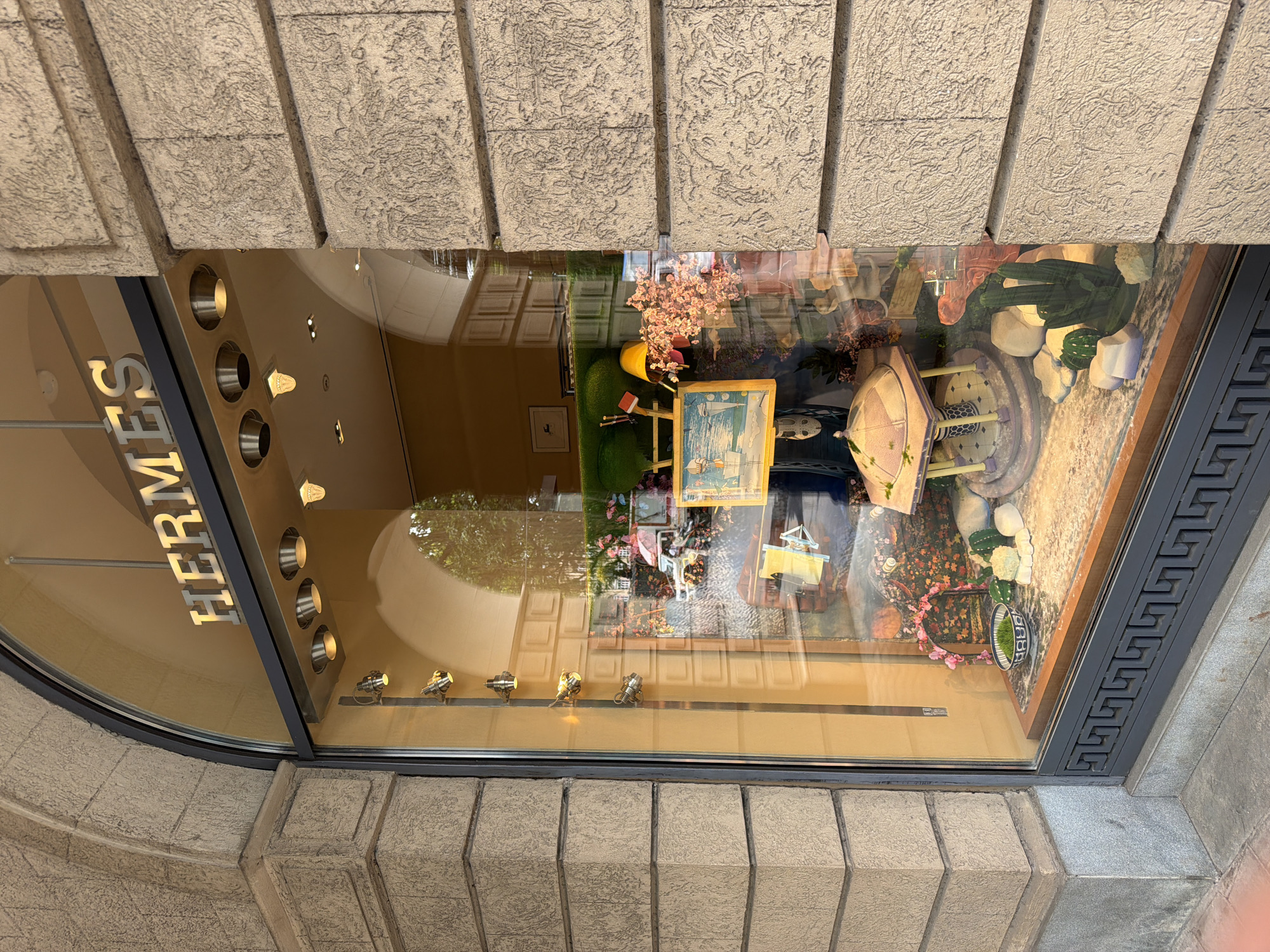 The image shows the exterior of an Hermès store, likely in Mumbai, India, given the architectural style and warm lighting indicative of a commercial establishment in the region. The store window features a display with several objects, including a small gazebo-like structure, a framed painting, floral arrangements, and what appears to be a small dish of green moss. The Hermès logo is prominently displayed above the window. Reflections in the glass reveal trees, a street scene with vehicles, and other buildings, suggesting an urban setting. The building's facade is made of textured stone blocks.