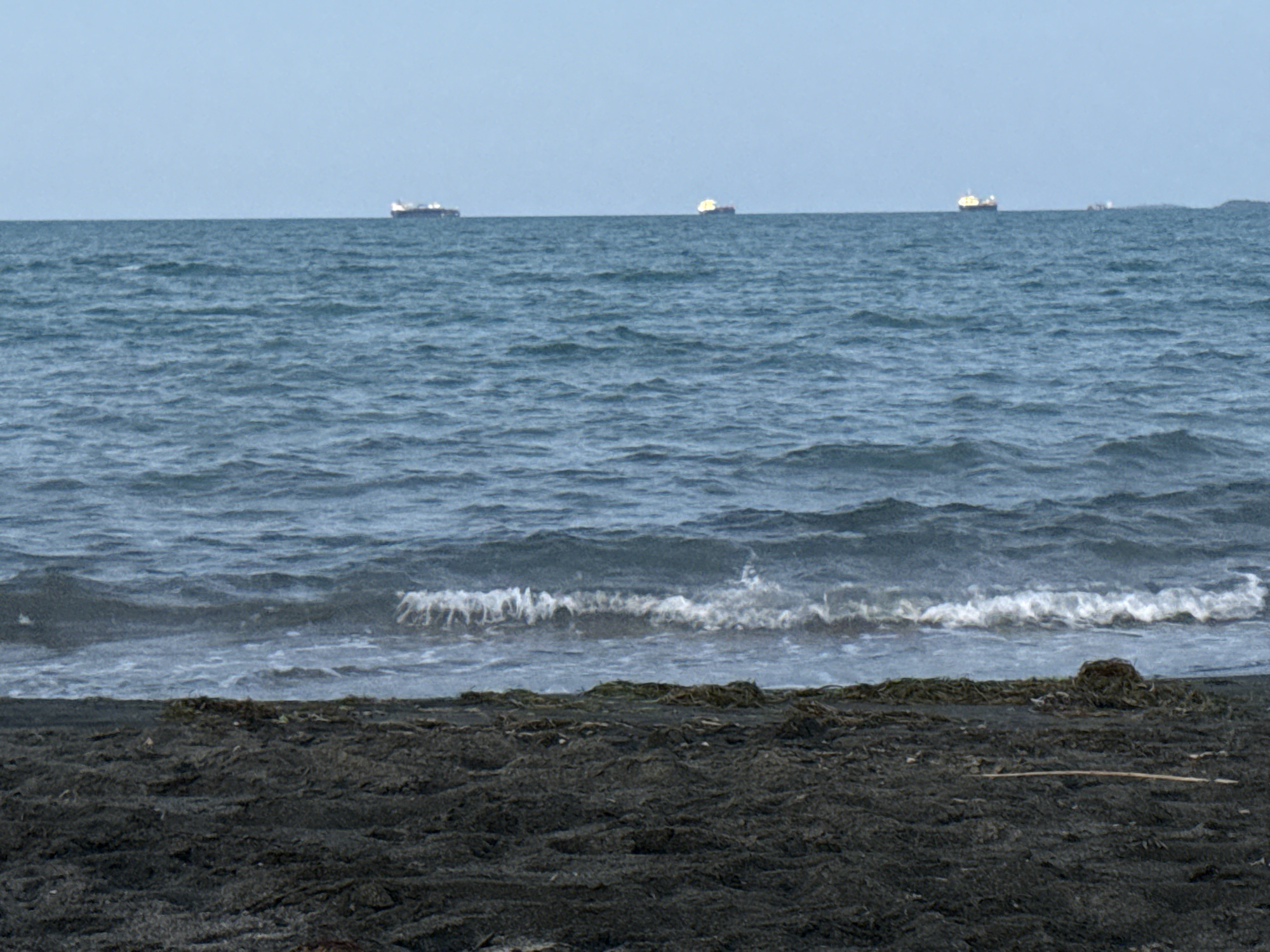 A wide, horizontal, medium-distance shot shows a dark, textured, sandy beach in the foreground with debris scattered across its surface. The middle ground consists of the dark blue sea with gentle waves and whitecaps, extending towards the horizon. The background is a clear, pale blue sky. Several large cargo ships are visible on the horizon, scattered across the sea. The image appears to be taken at the coast of Burgas, Bulgaria, with a focus on the natural elements of the beach and the sea, along with the presence of maritime activity.