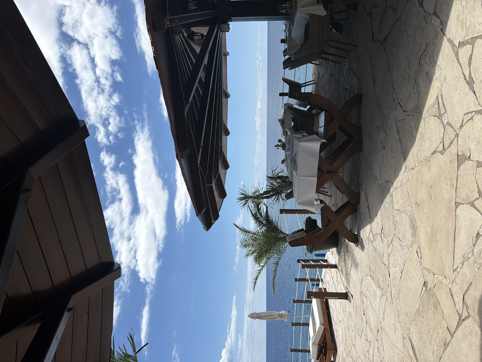 This is a photograph of an outdoor restaurant patio overlooking the sea, likely in Novoselje, Montenegro. The patio is paved with large, irregular light-colored stones and is partially covered by a dark wooden structure with a sloped roof.
In the foreground, two wooden chairs with an 'X' shaped base are positioned around a table covered with a white tablecloth. The table is set with place settings and condiments. To the left of this table, another table is partially visible, also set with a white tablecloth.
Further back, beyond the tables, there is a railing separating the patio from the water. Beyond the railing, a white beach umbrella is partially opened, and a wooden lounge chair with white cushions is set up. Two palm trees are visible on the patio, one in a large dark pot.
The sea is a deep blue, meeting a bright blue sky with scattered white clouds. The horizon is clearly defined between the sea and sky. The image captures a serene, coastal dining setting. There are no people visible in the image.