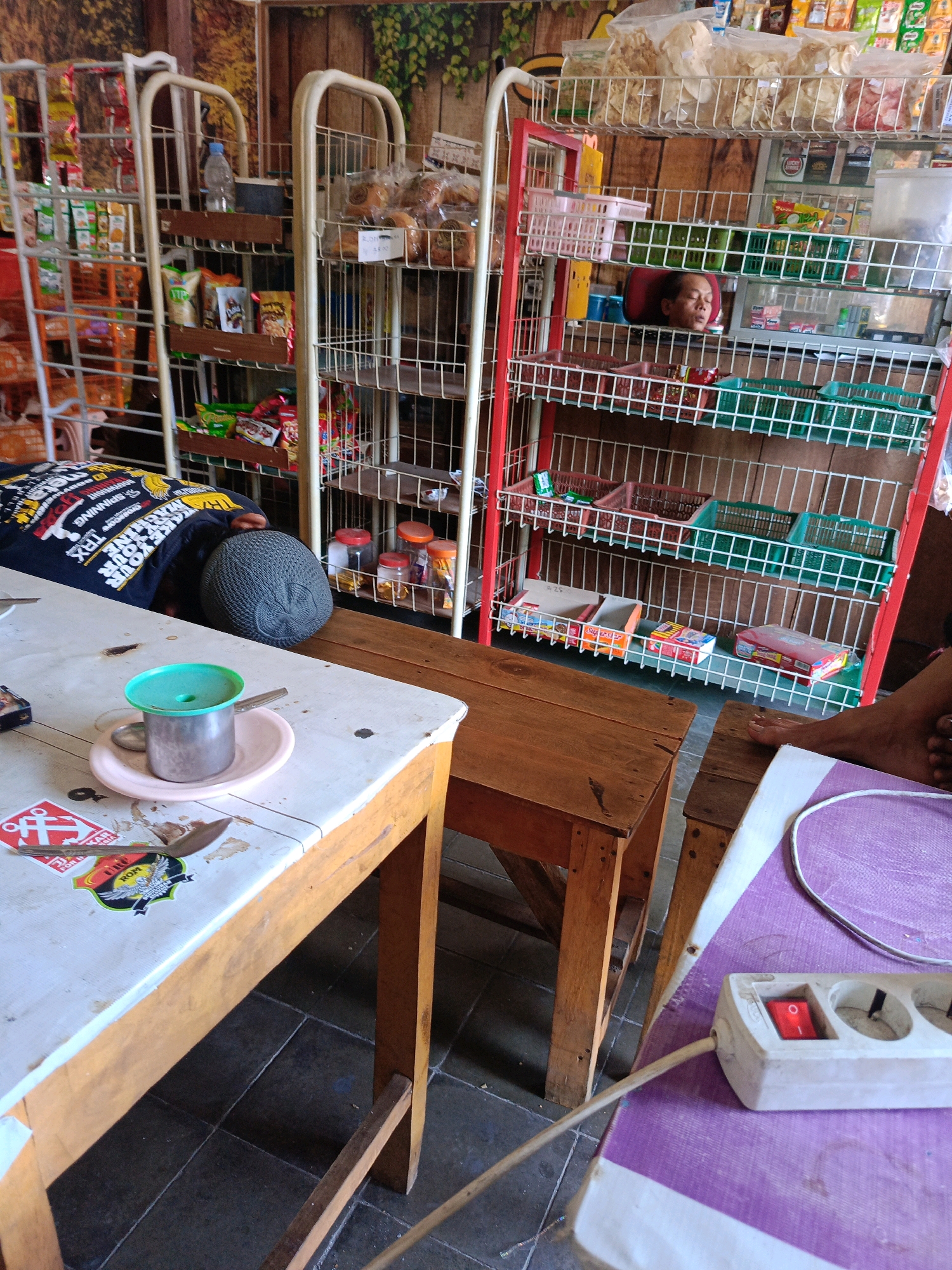 A man is sleeping on a table at a shop in Kebonagung, Indonesia. He is wearing a black t-shirt with white text and a grey skull cap. A metal cup with a green lid sits on a pink saucer in front of him on the table. There are stickers on the table, and a spoon is next to the cup. Behind the man, shelves are filled with various items including snacks, bottles, and baskets. To the right of the shelves, a power strip with a red switch is visible on a purple surface. The floor is tiled.