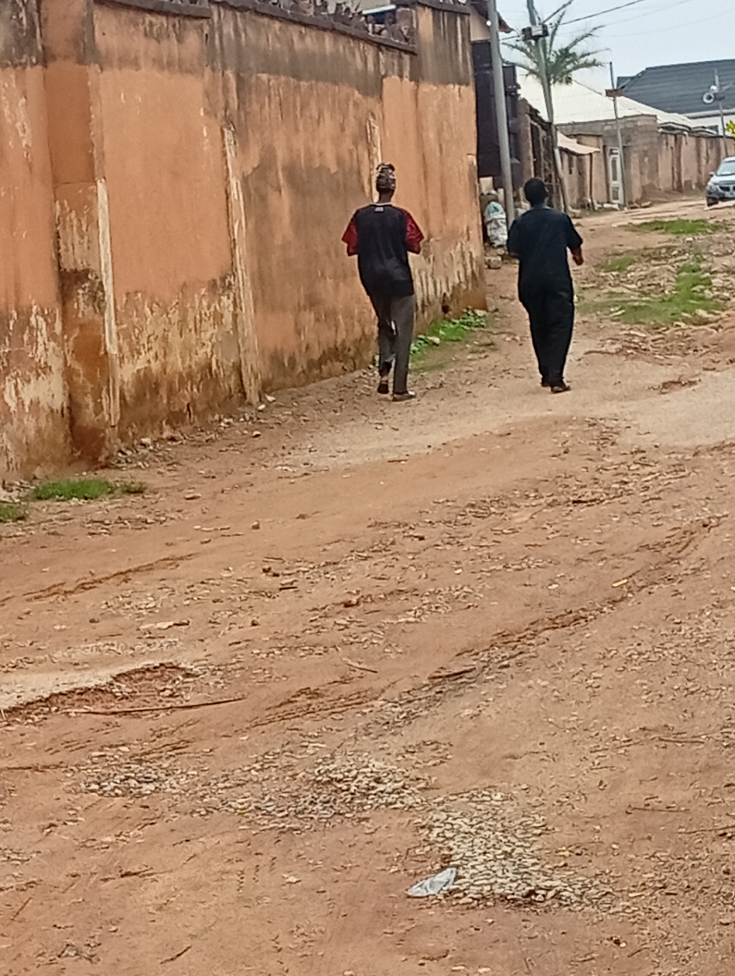 Two individuals are walking away from the camera on a dirt road in Rafin Par, Nigeria. The person in the foreground on the right wears a black outfit. The person ahead on the left wears a black t-shirt with red accents and grey pants. They are walking past a dilapidated, light brown wall. The environment appears to be a rural or semi-urban area, with sparse vegetation and other buildings visible in the background. A car is parked further down the road on the right. The road surface is uneven and covered with dirt and small stones.