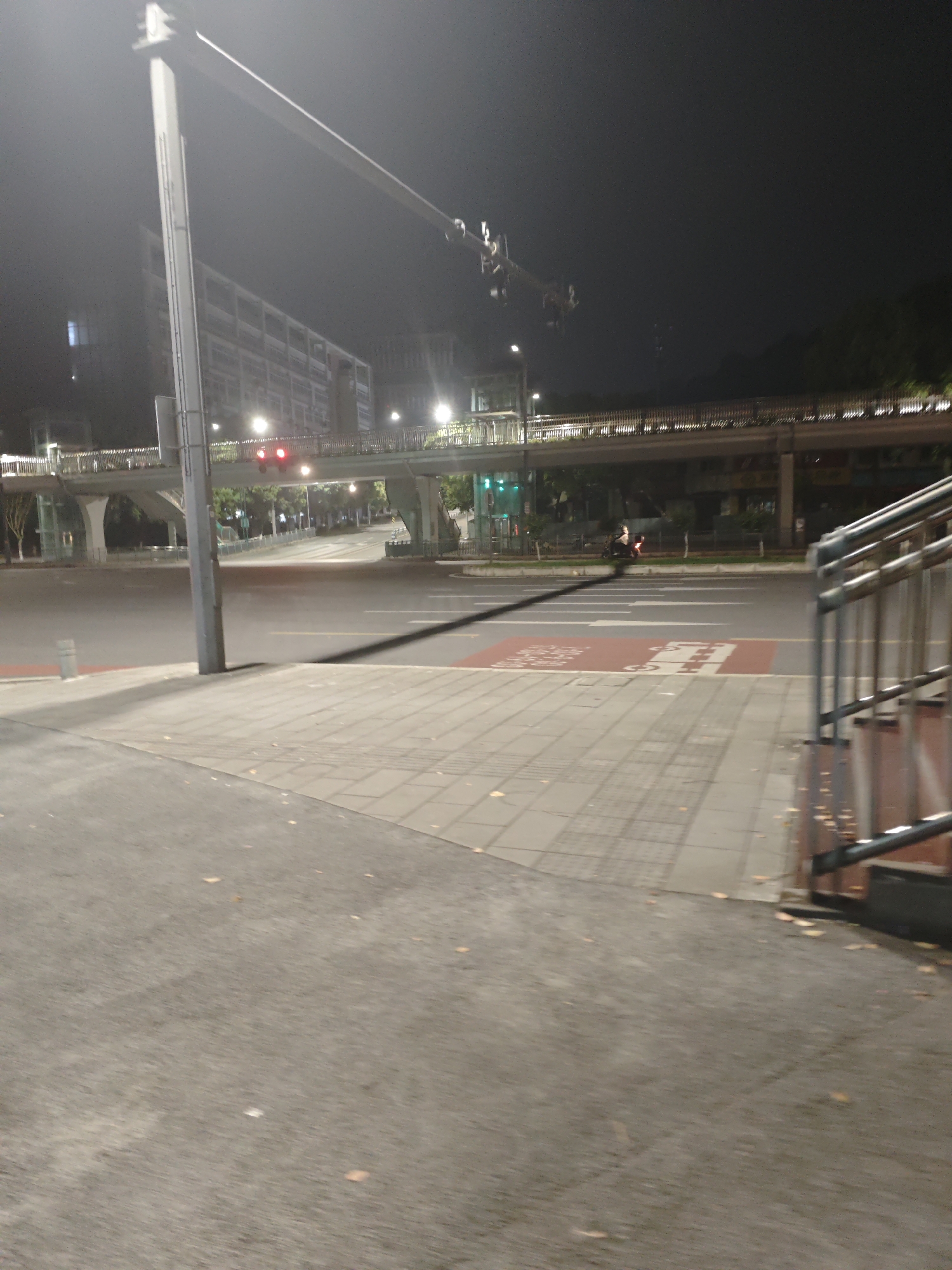 The image depicts a street scene at night in Chongqing High-tech Zone, China. A pedestrian walkway spans the road, supported by concrete pillars. Red traffic lights are visible on the walkway, while green lights are seen on the road below. A scooter with two occupants is on the road. The foreground is a sidewalk paved with gray bricks. A metal railing is partially visible on the right side of the image.