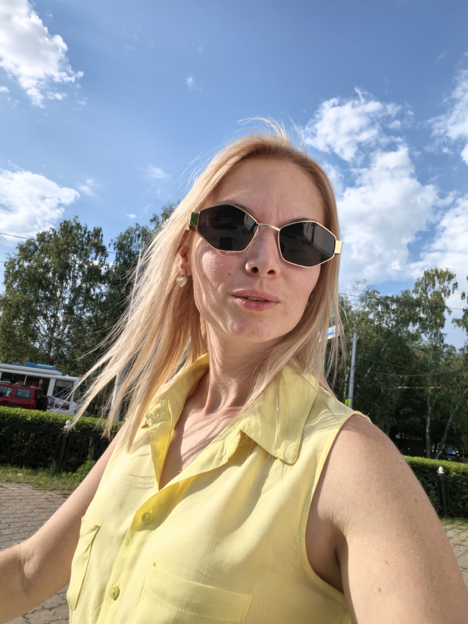 A Caucasian woman with blonde hair and sunglasses is captured in a selfie outdoors, under a bright blue sky with scattered white clouds. She is wearing a sleeveless yellow collared shirt. The background includes green trees, a paved walkway, and some vehicles, including a red car and what appears to be a public transport vehicle, possibly a tram or bus, with a white and blue exterior. The scene suggests a sunny day in an urban or park-like setting within the city of Sterlitamak, Russia.